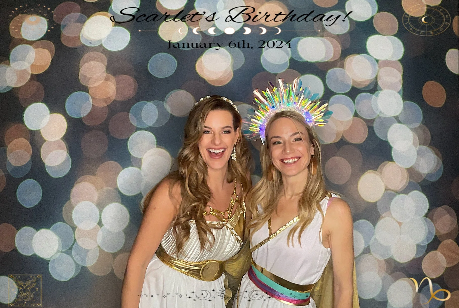 Two women in Greek-themed costumes celebrating Scarlet's birthday, January 6th, 2024, standing in front of a colorful bokeh background, with one wearing a crown and the other a halo-like decoration.
