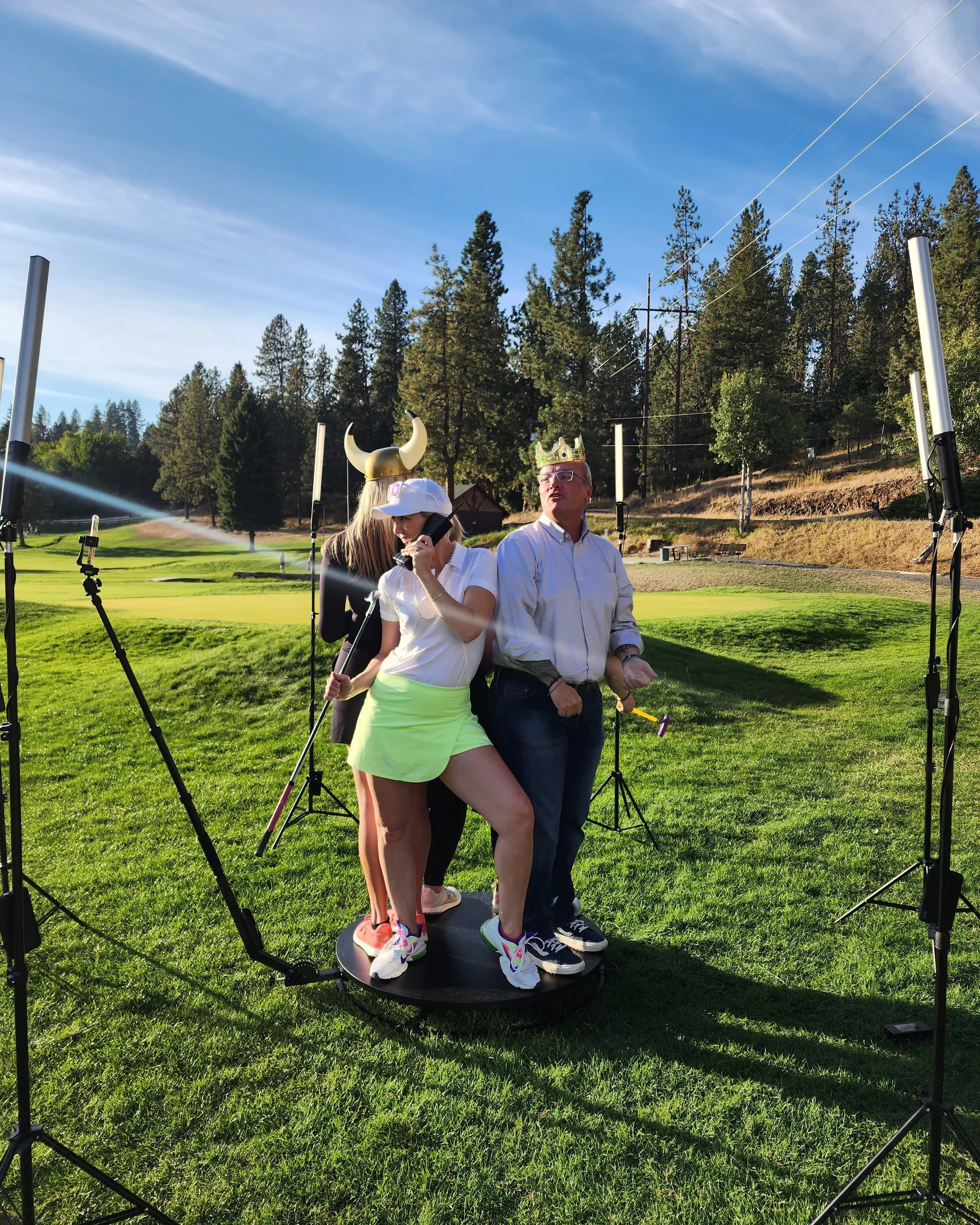 People wearing Viking hats standing on a rotating platform in a grassy golf course, surrounded by lighting equipment, with trees and a blue sky in the background.