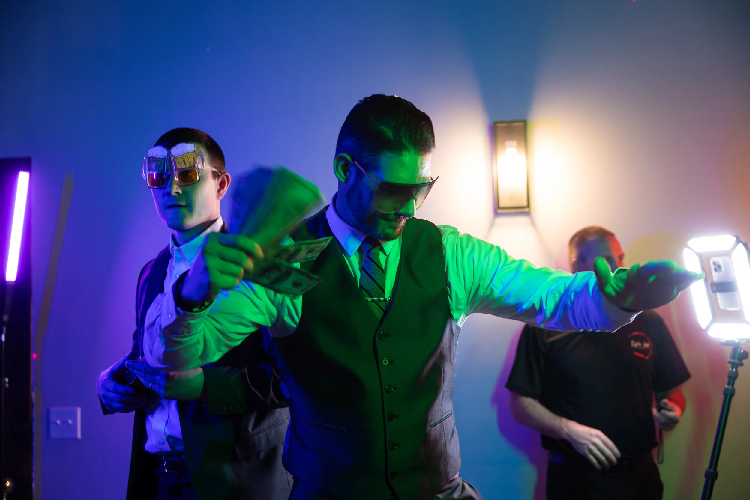 Party scene with three men, two wearing sunglasses, one holding cash, colorful lighting, and a lit room.