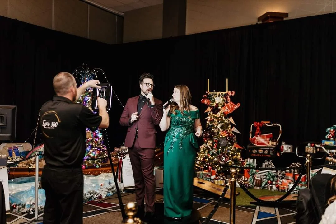 Two people with microphones speaking on stage decorated with Christmas trees and gifts, while a man records them with a camera.