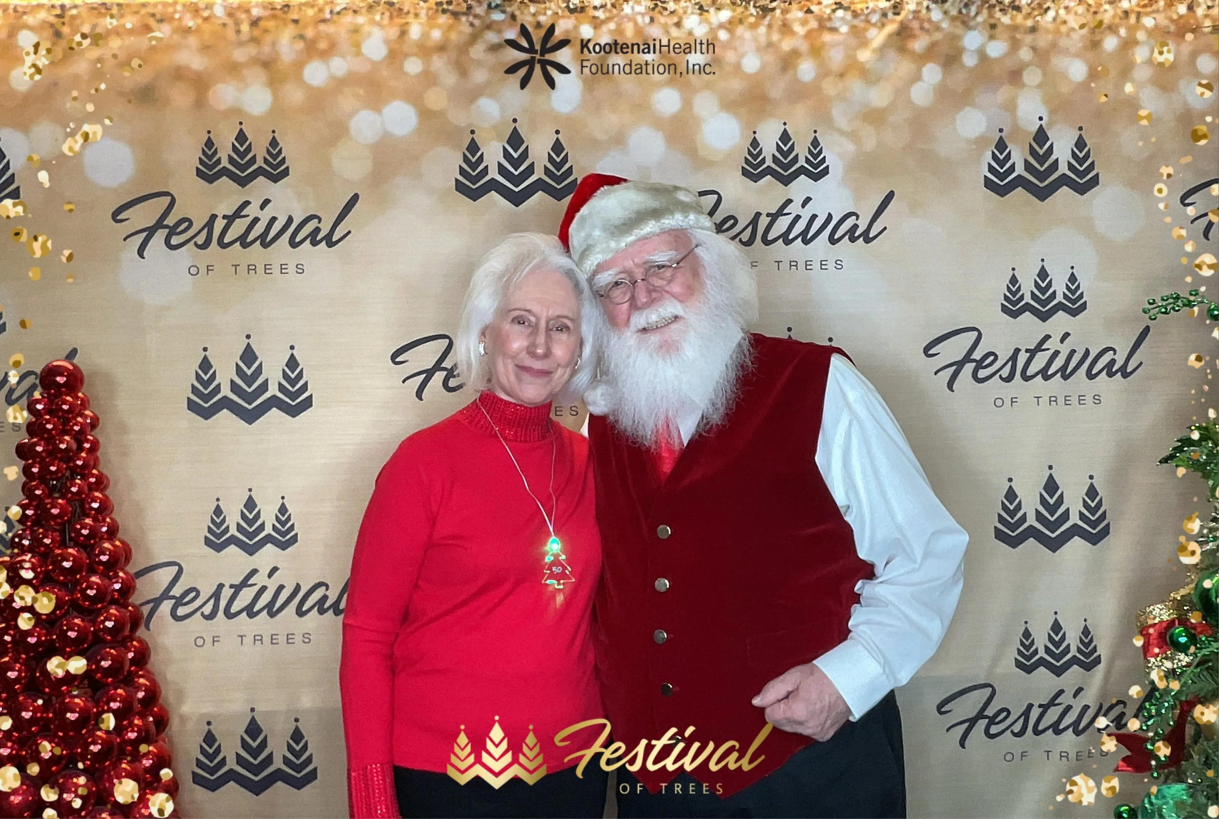A woman and man dressed as Santa Claus standing together at the Festival of Trees event, with a decorated Christmas tree on the left and festive background with the event's logo.