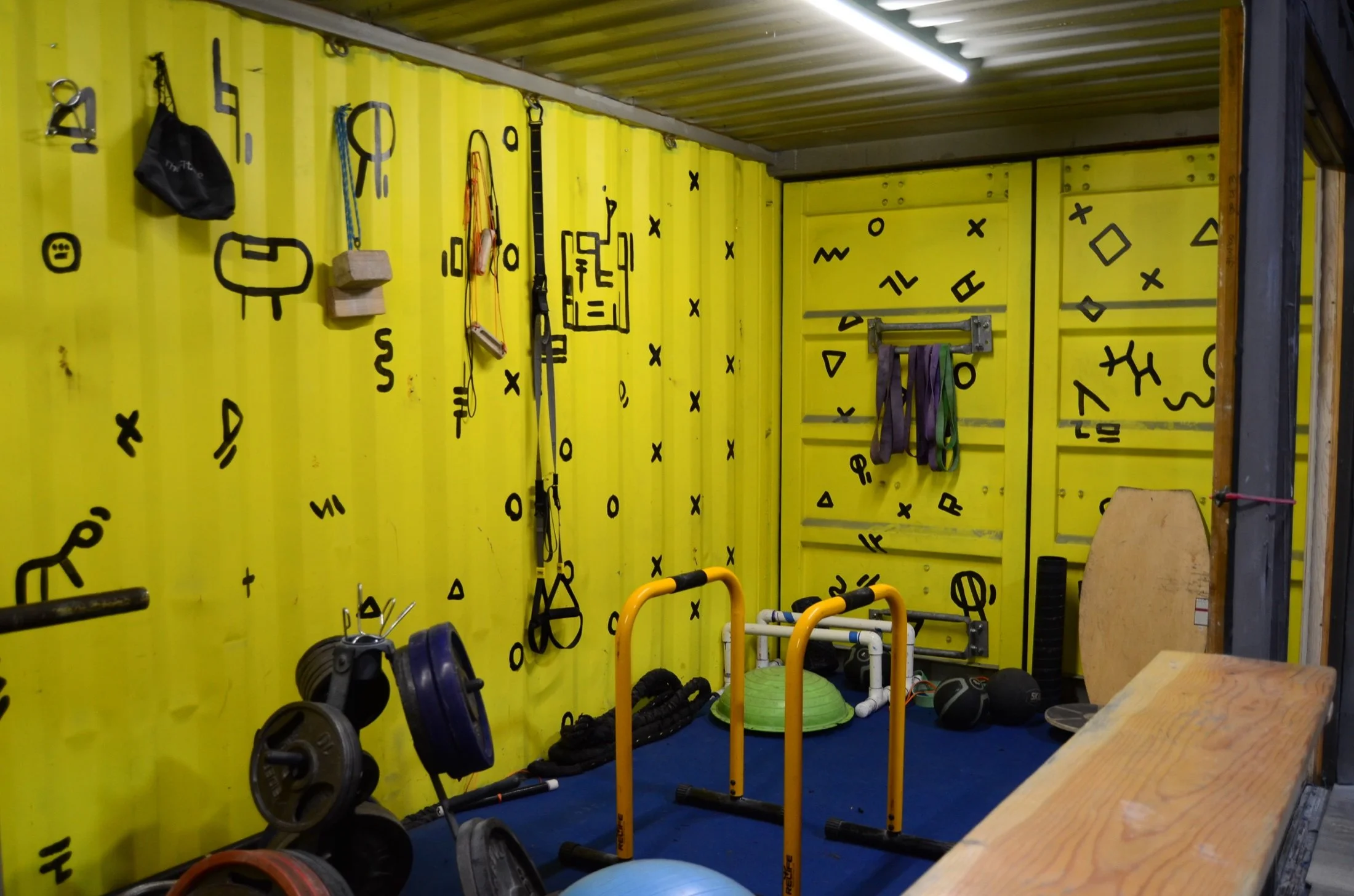 A gym corner with yellow walls decorated with black geometric and doodle patterns. Various workout equipment such as kettlebells, medicine balls, resistance bands, and weights are organized on the floor. A yellow squat rack with purple and green resi