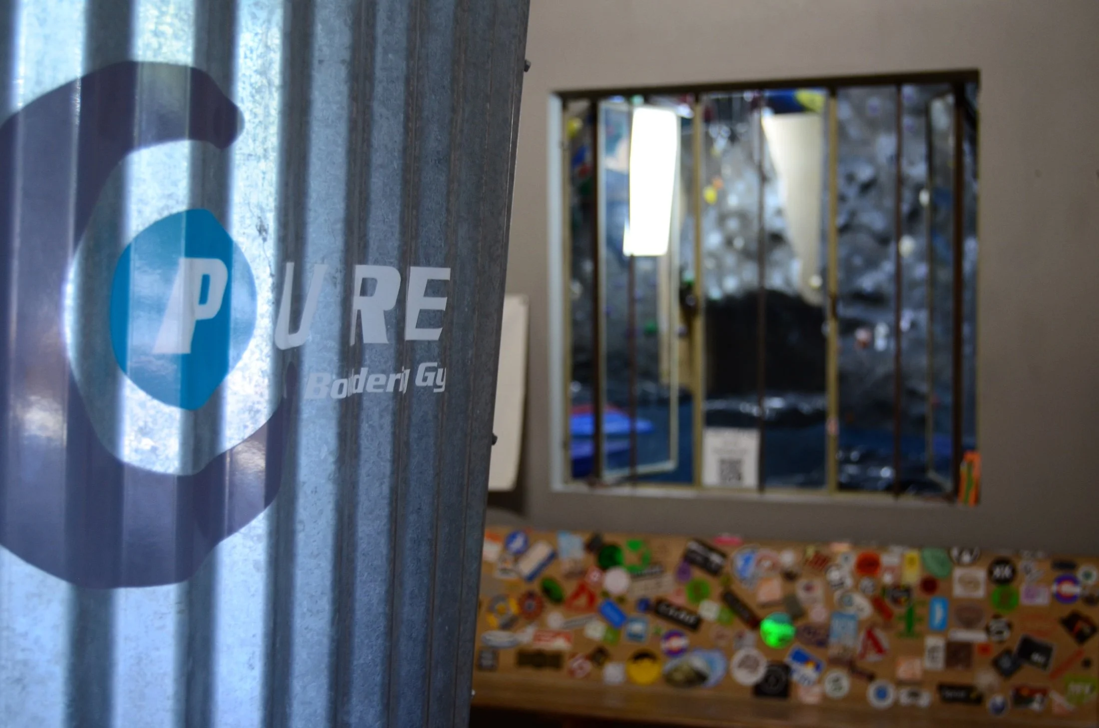 Close-up of a metallic pole with a blue and white sticker reading 'PURE' and 'Bolder G' partially visible. In the background, a mirror reflects a cluttered room with various objects, including colorful stickers or pins, and a black object on a counte