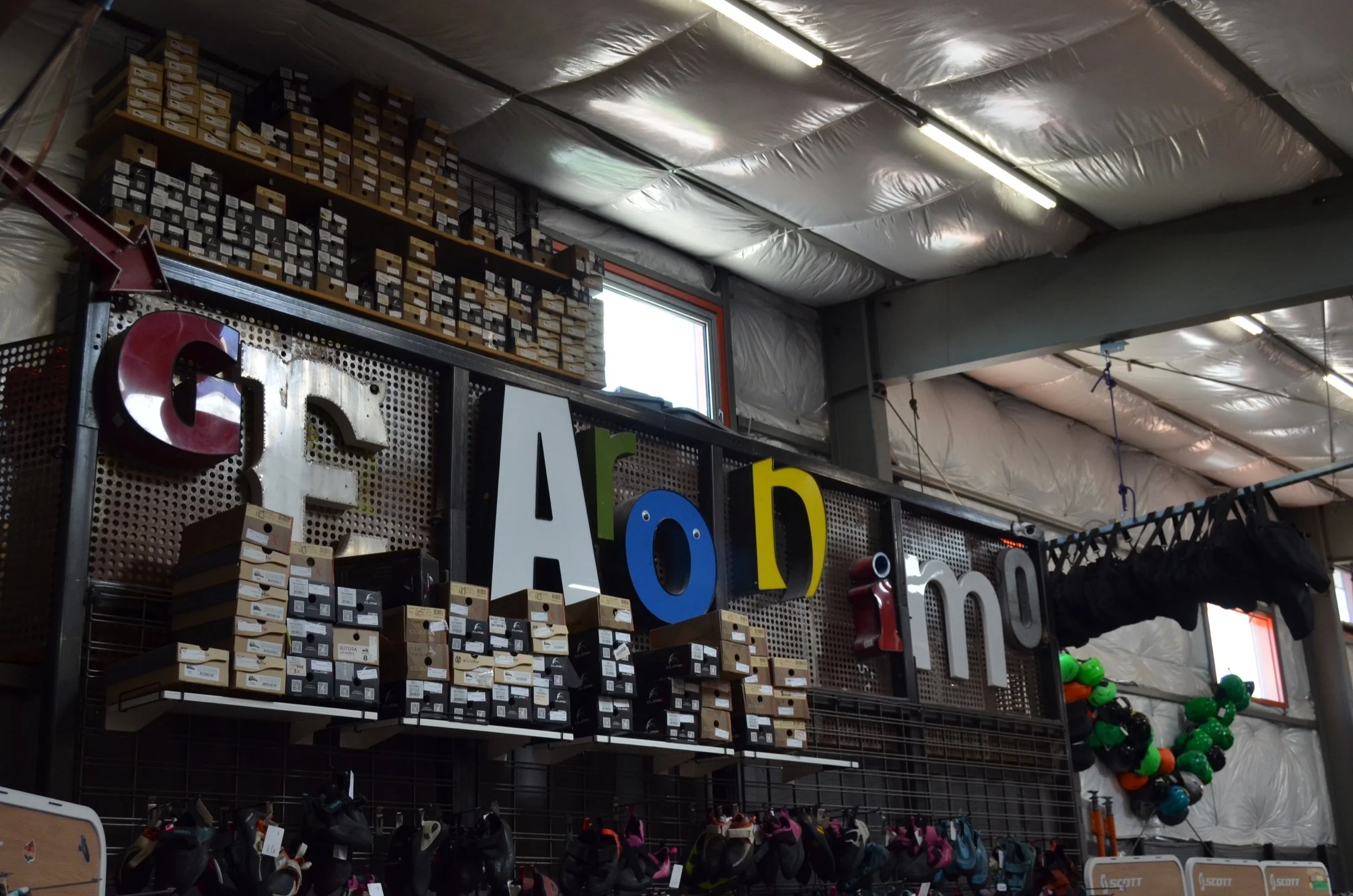 Interior of a sporting goods store with shelves stocked with boxes of footwear, colorful helmets, and equipment, along with some rack-mounted shoes and signs in front.