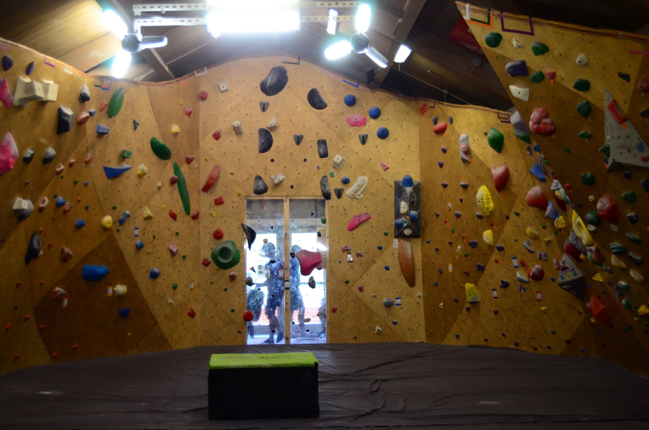 An indoor rock climbing gym with yellow walls covered in various colorful climbing holds. There are bright lights and ceiling fans visible in the ceiling, and a person can be seen climbing through the window at the back.