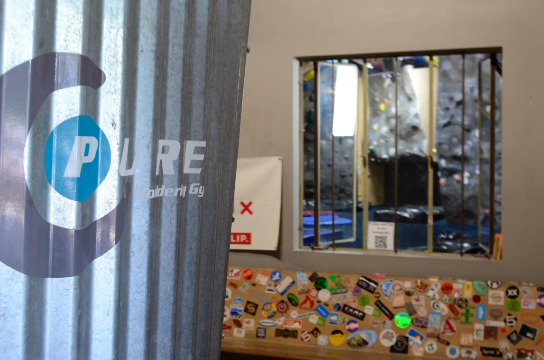 A metal sign with a blue and white logo for Pureleds, partially overlapping a room with a barred window and a nearby wall covered in stickers. The room appears to be a rock climbing gym, with climbing holds visible through the window.
