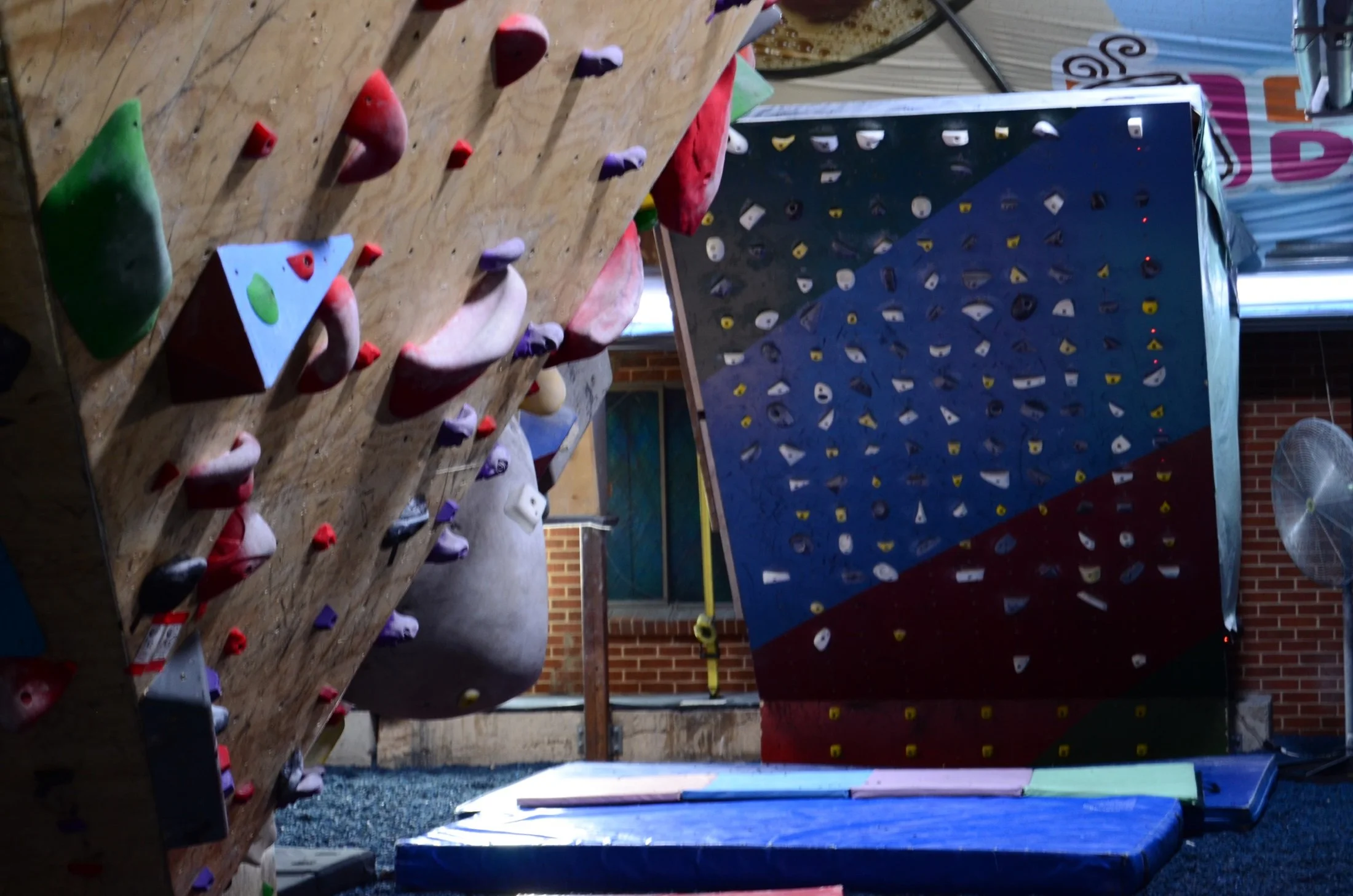 Indoor rock climbing gym with a climbing wall featuring various holds and volumes in different colors, and a separate bouldering section with a taller wall.