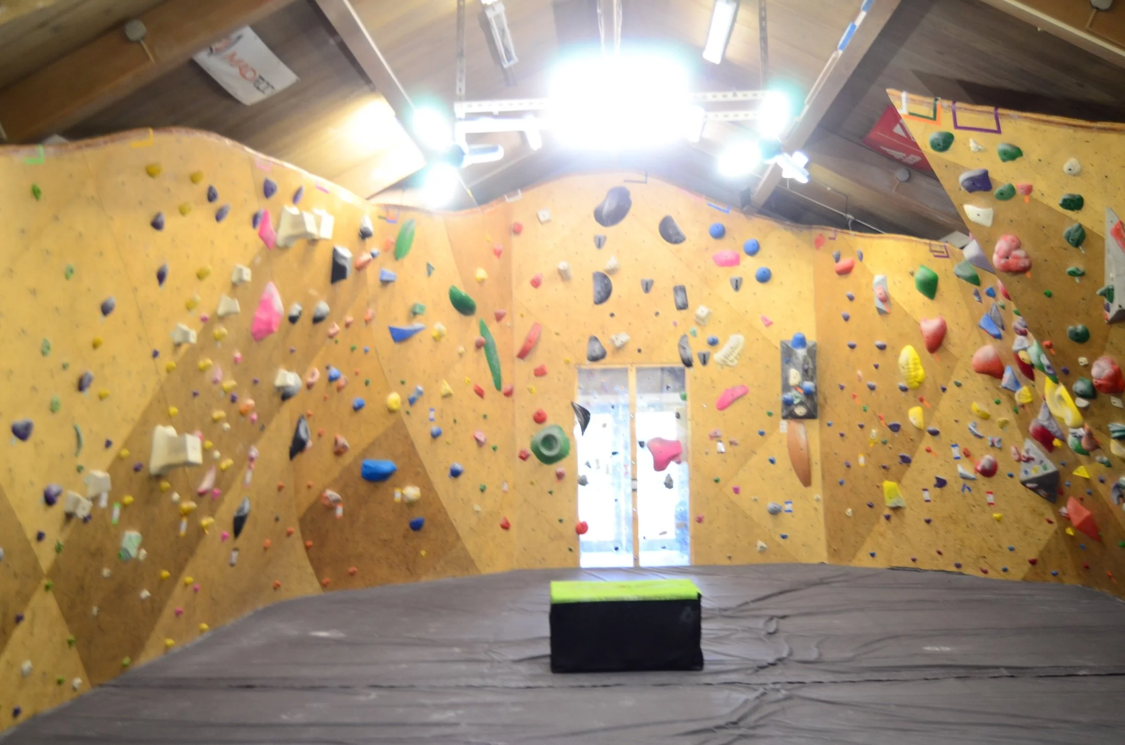Indoor rock climbing gym with a large climbing wall covered in colorful holds, illuminated by bright ceiling lights, with a padded floor at the bottom for safety.