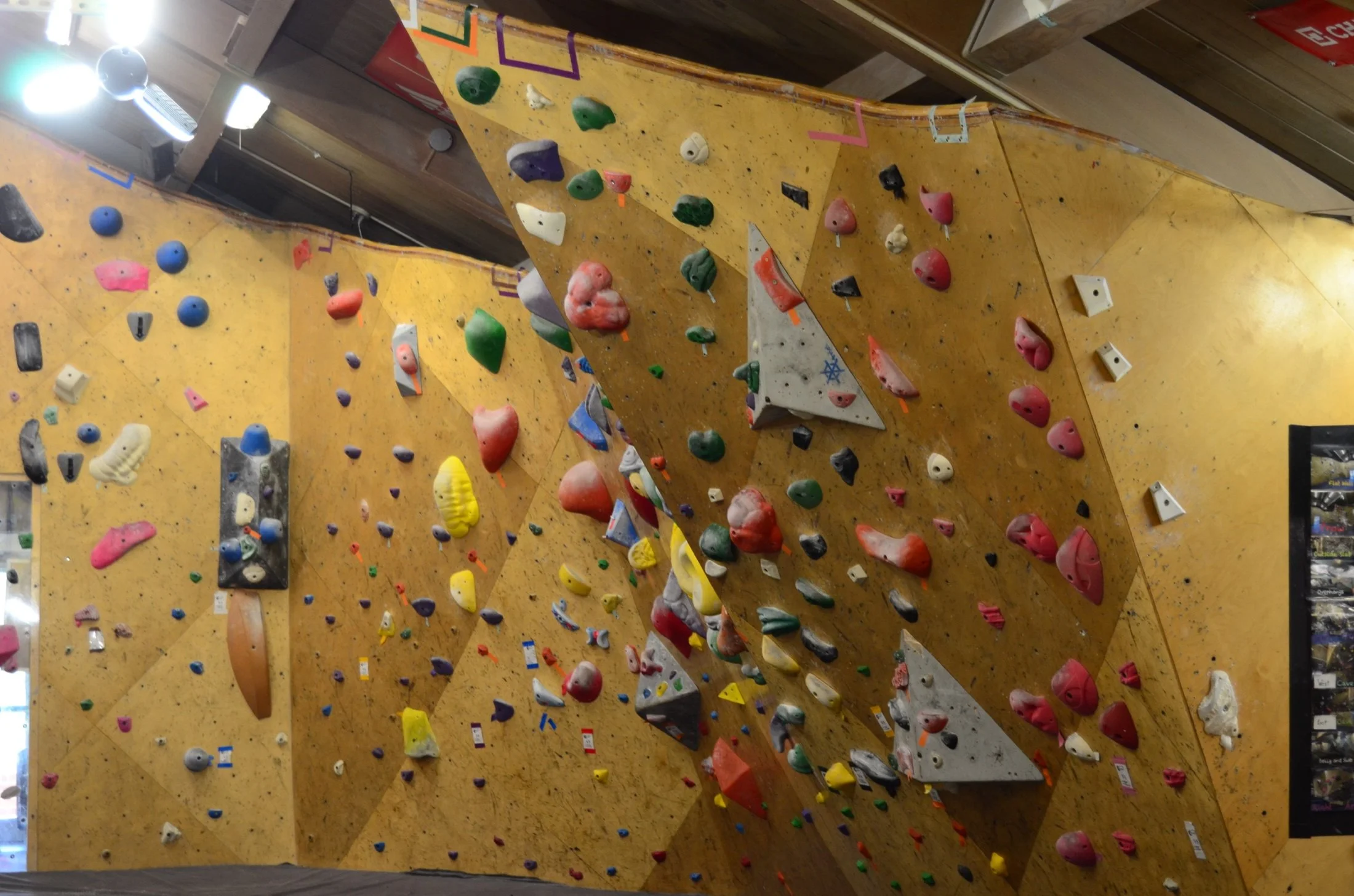 An indoor rock climbing gym with yellow climbing walls covered in various colorful holds and grips. The walls are angled with some sections overhanging. Bright lights illuminate the area.