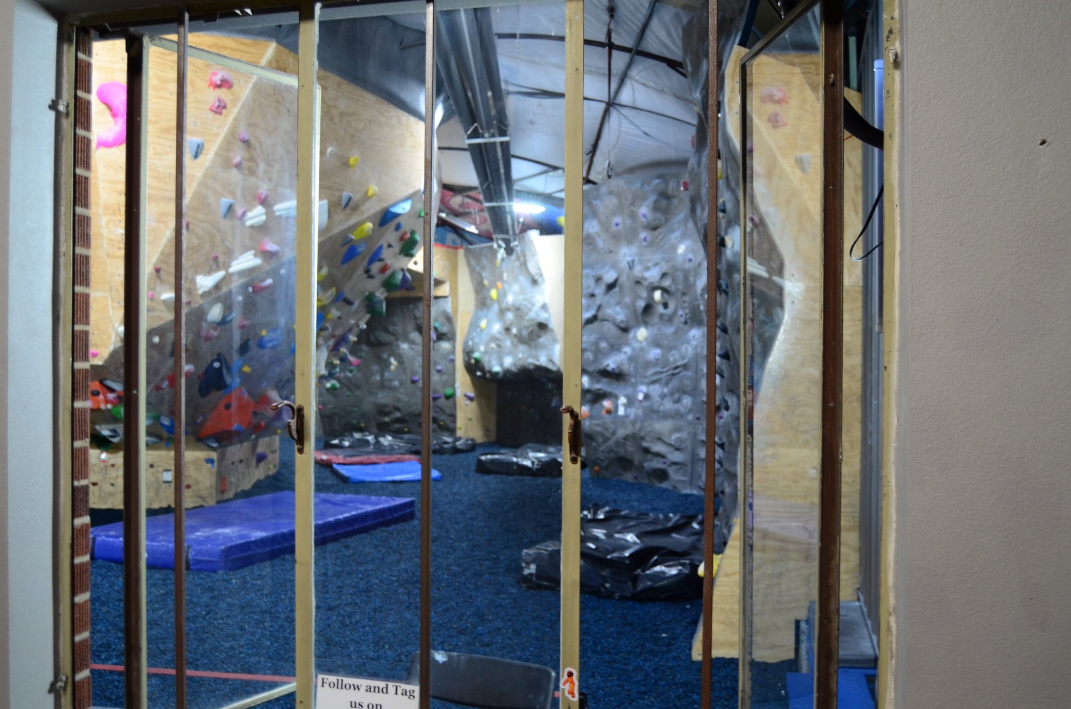Indoor rock climbing gym seen through a metal frame window with a sign that says 'Follow and Tag us on' at the bottom. The climbing area contains various colorful holds and a wall with different angles, including a steep overhang.