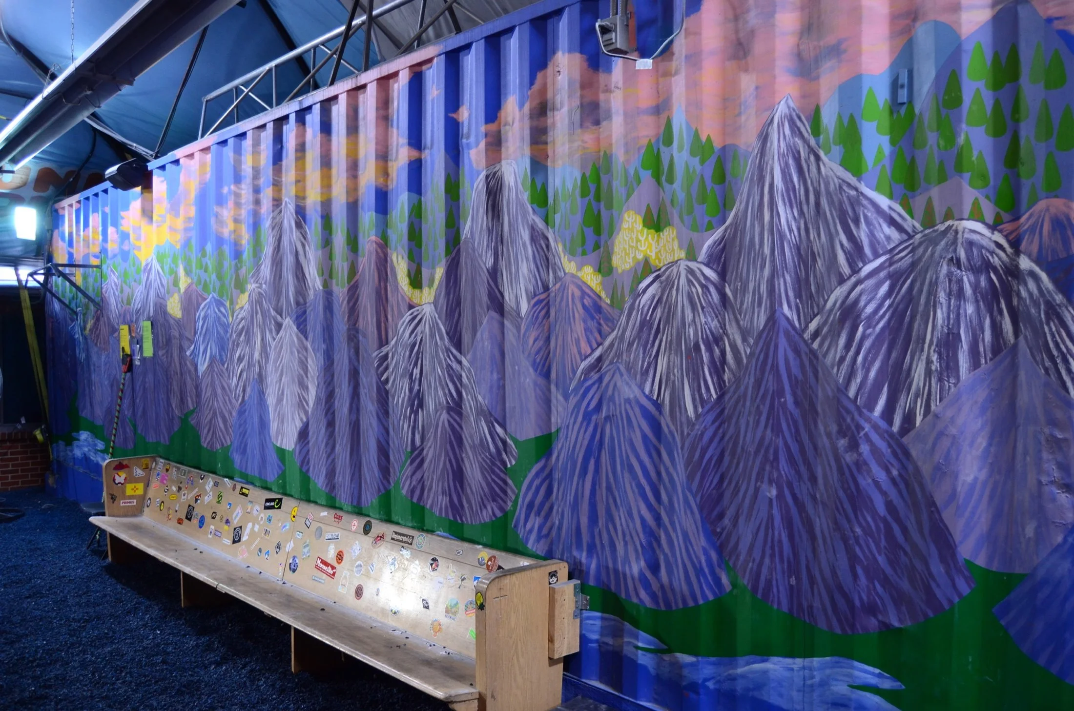 Colorful mural of mountains, trees, and a sunset sky with clouds painted on a wall, with a wooden bench and a sticker-covered board in the foreground.