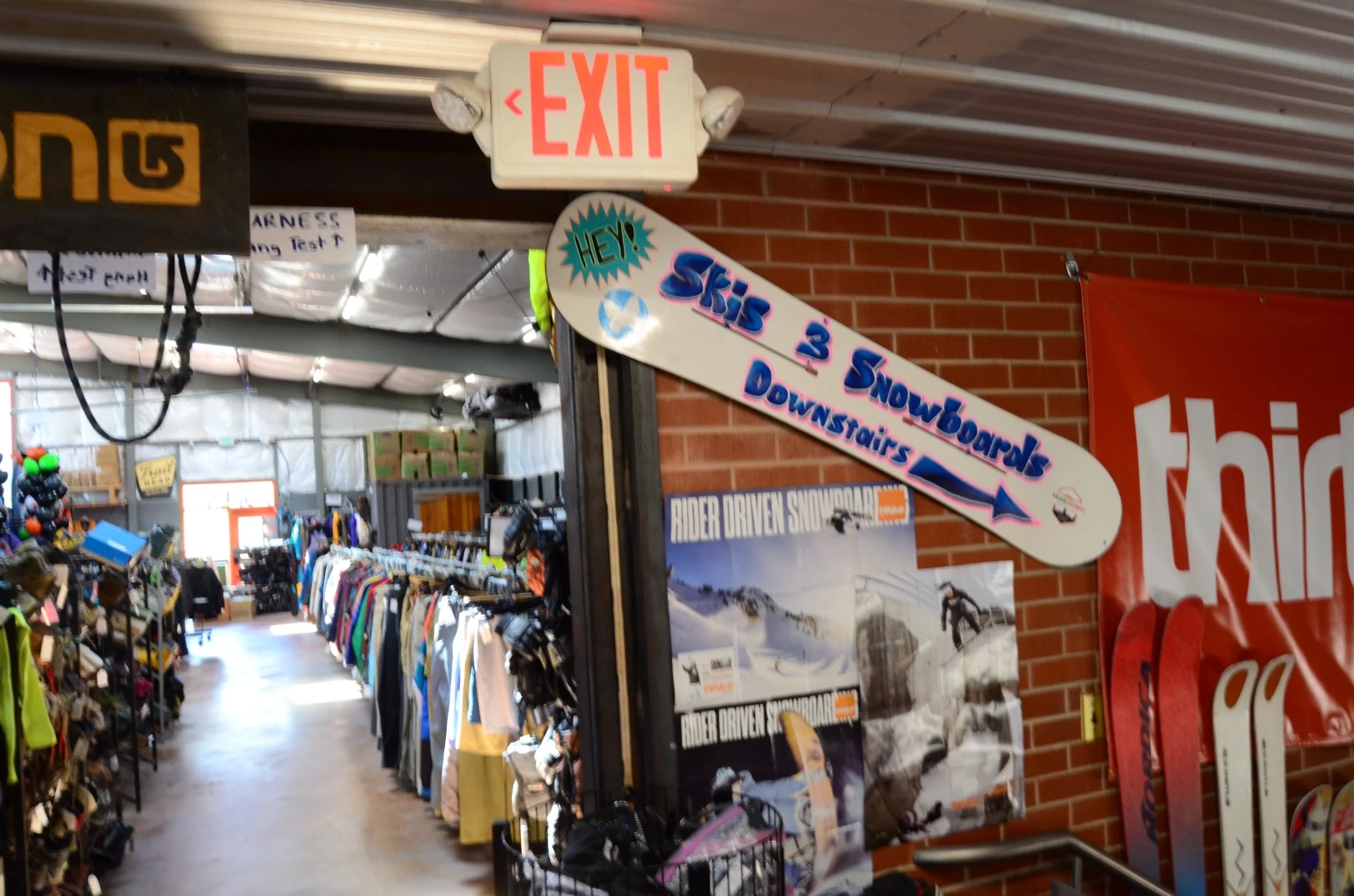 Interior of a sporting goods store with clothing racks, signs for snowboarding, and an orange brick wall.