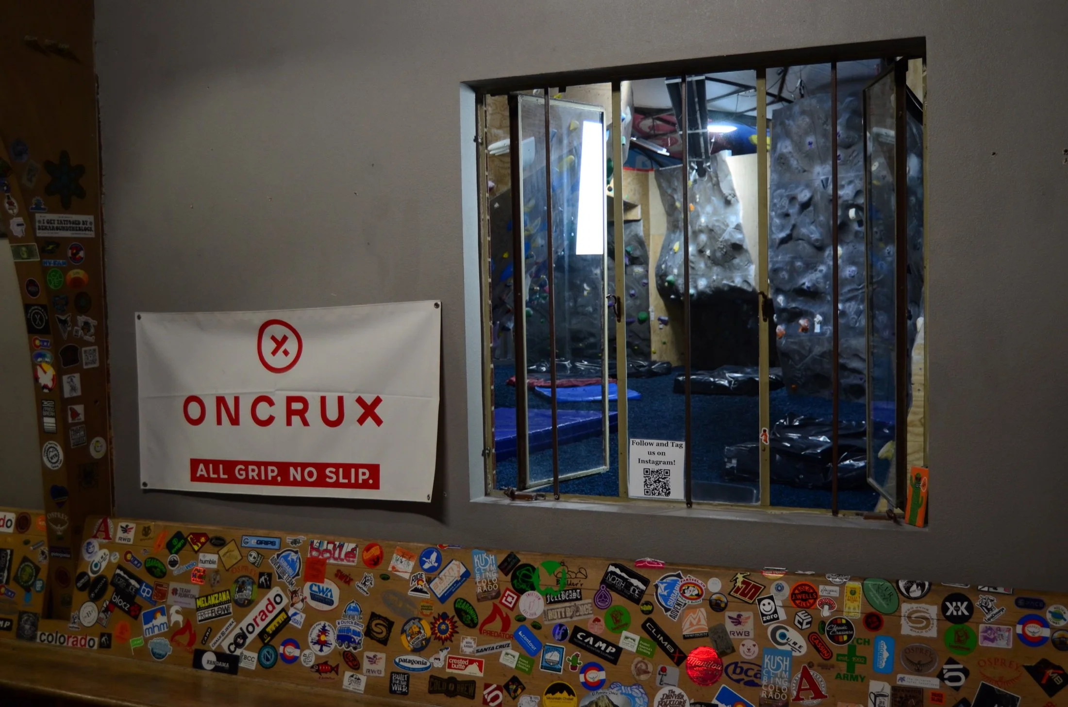 Indoor rock climbing gym viewed through a window with a security grill. The gym has climbing walls with holds and mats on the floor. You can see a sign that says 'Oncrux, All Grip, No Slip' and a QR code below it. The window is surrounded by colorful