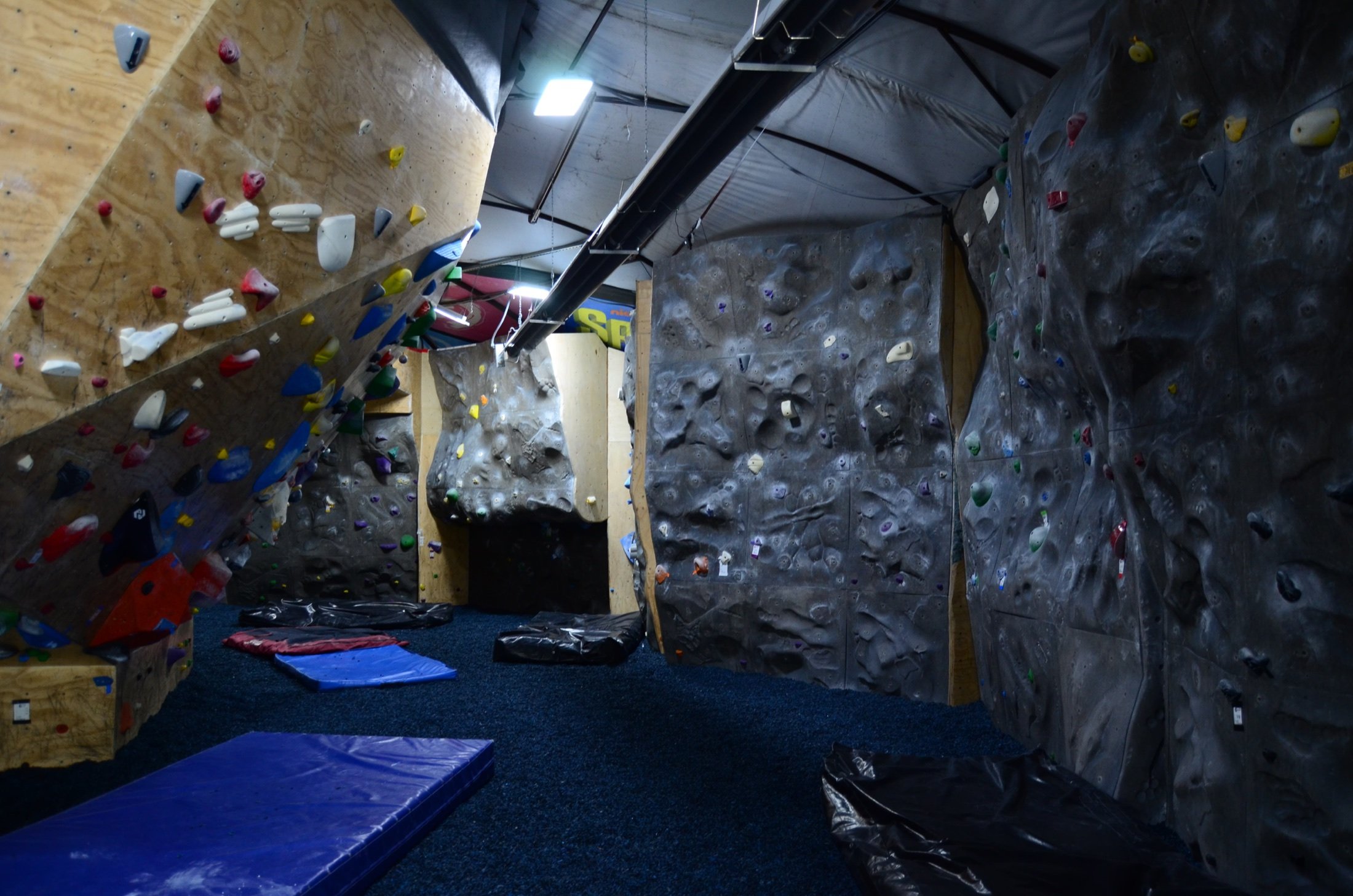 Indoor bouldering gym with climbing walls covered in colorful holds, padded mats on the floor, and climbing chalk bags placed nearby.