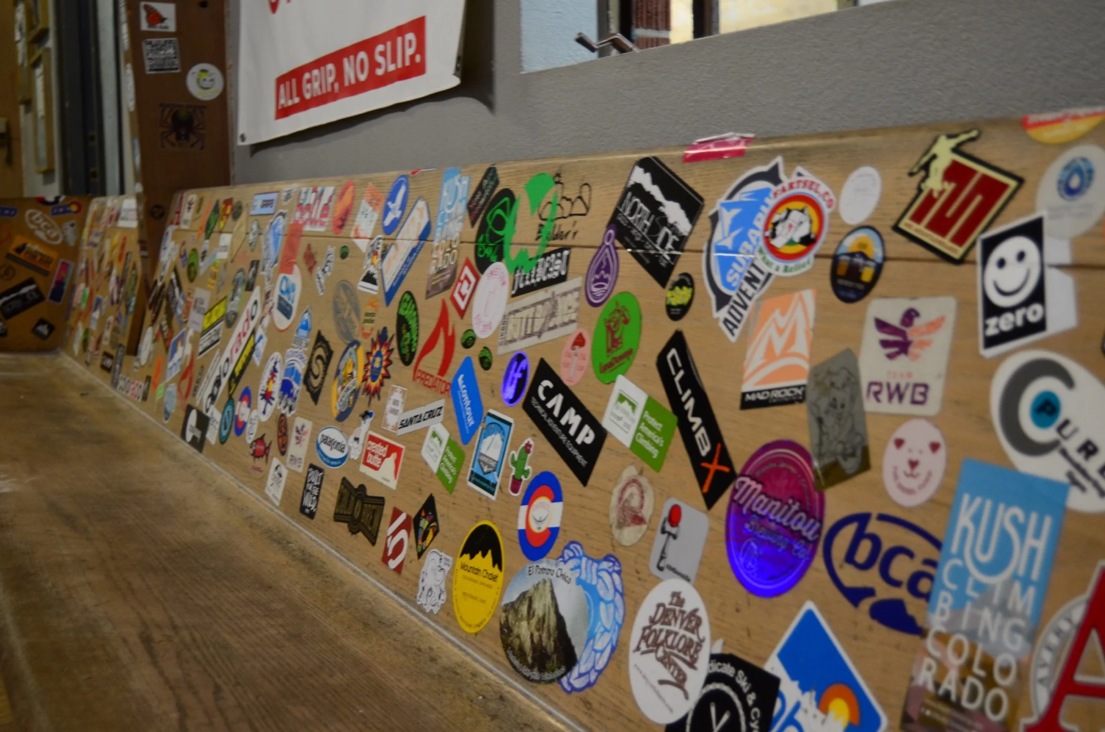A wooden surface covered with various stickers and decals, with a sign that reads 'ALL GRIP, NO SLIP' in the background.