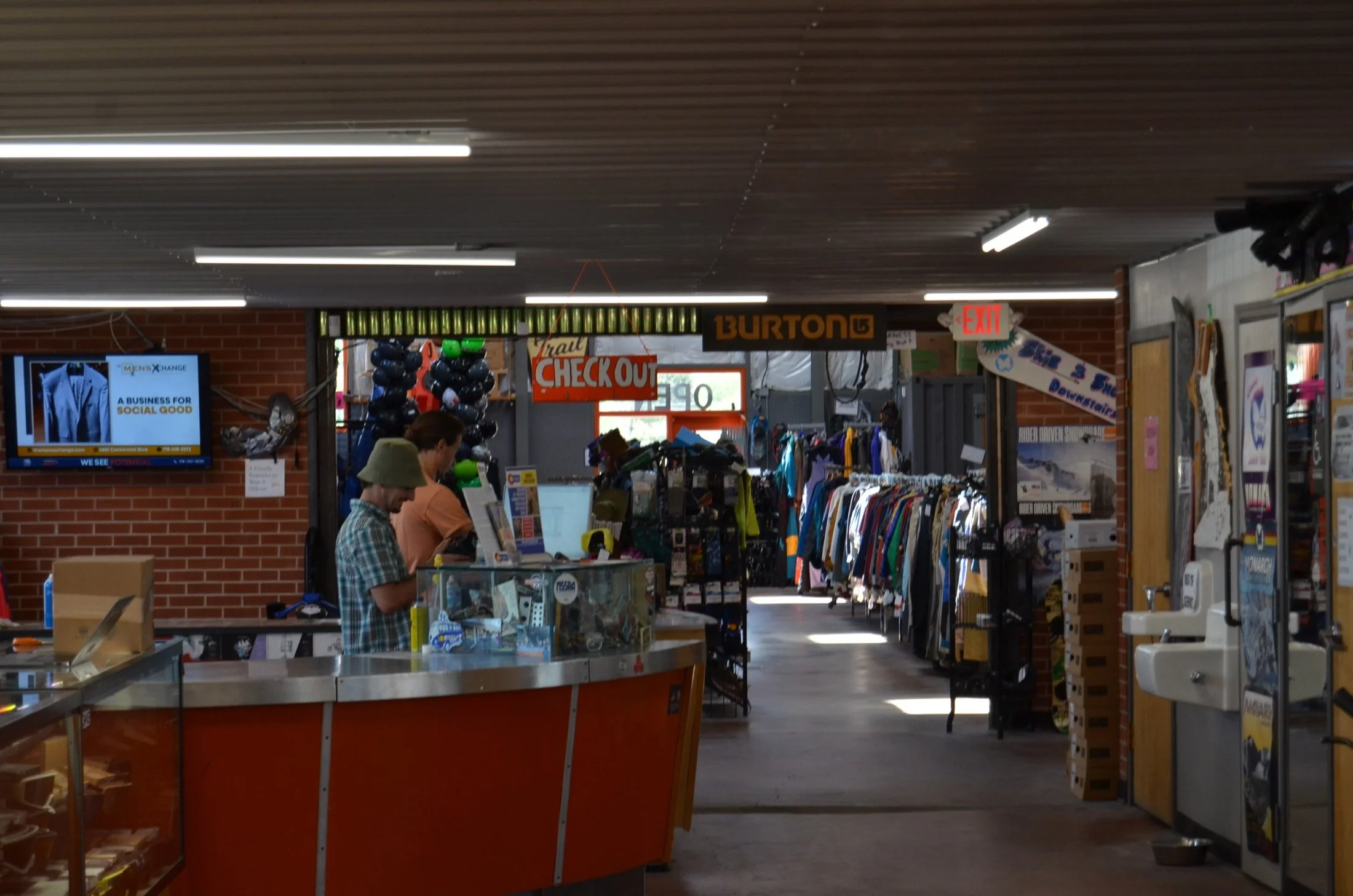 Inside a retail store with clothing racks, a checkout counter, and a digital screen, with customers shopping and the store's ceiling lights visible.