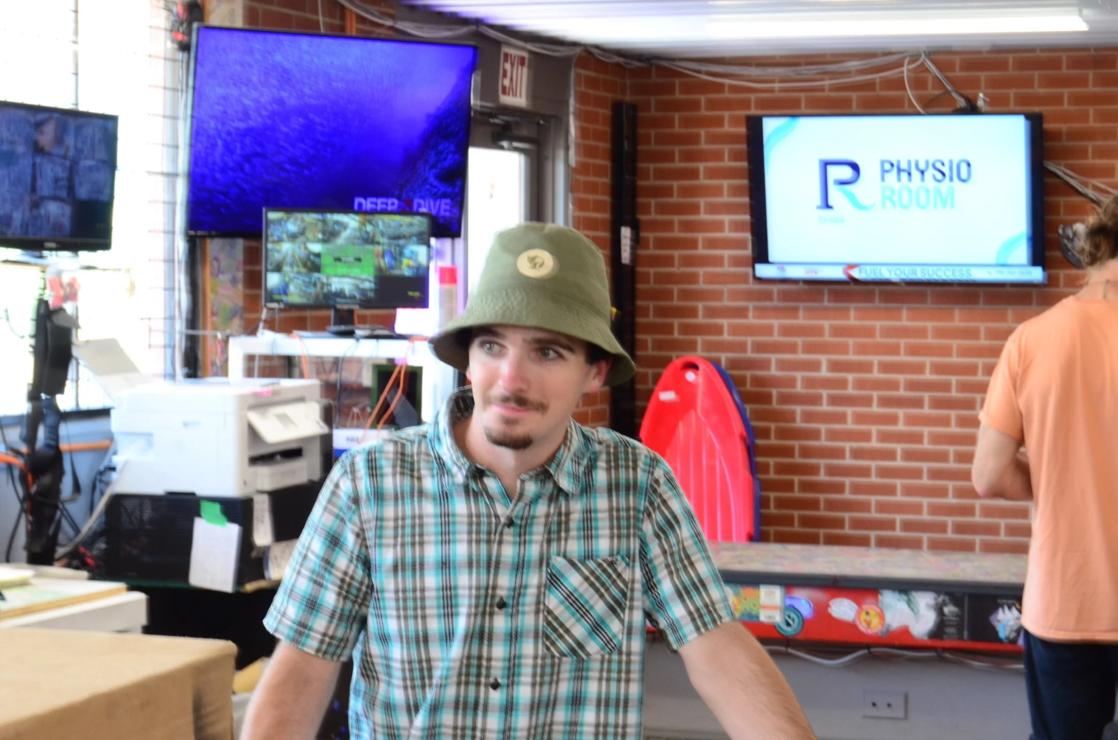 A young man with a beard, plaid shirt, and bucket hat sitting at a desk inside a room with brick walls. There are three screens on the wall behind him, with logos that say "Deep Dive" and "Physio Room." Other people are present, and various equipment