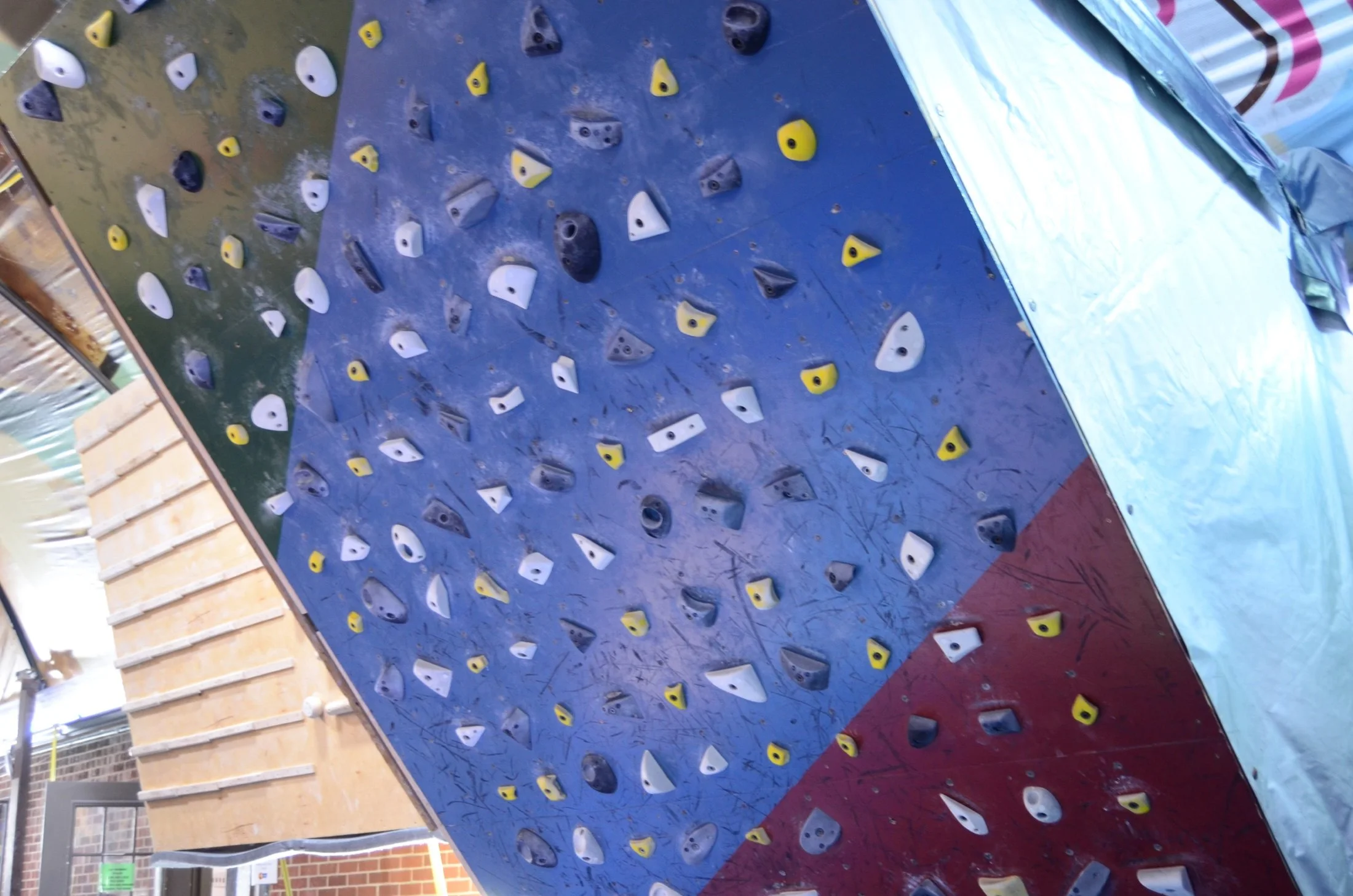 Indoor rock climbing wall with multicolored holds on blue, yellow, black, gray, and white surfaces.