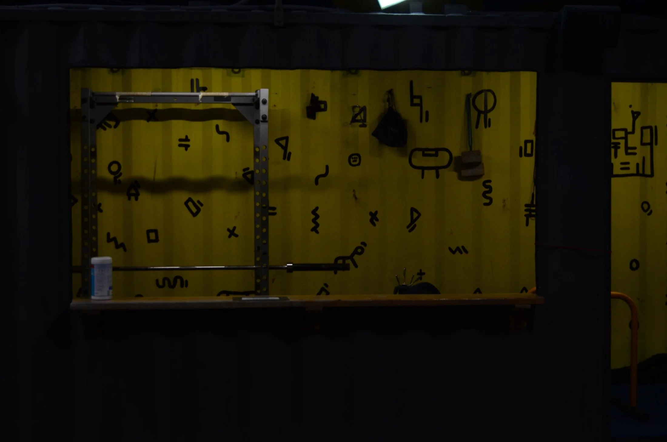 A yellow wall with black symbols and letters, a metal rack on the left, a black bag hanging, and a small wooden block on the right. Part of a can or container is visible at the bottom left.