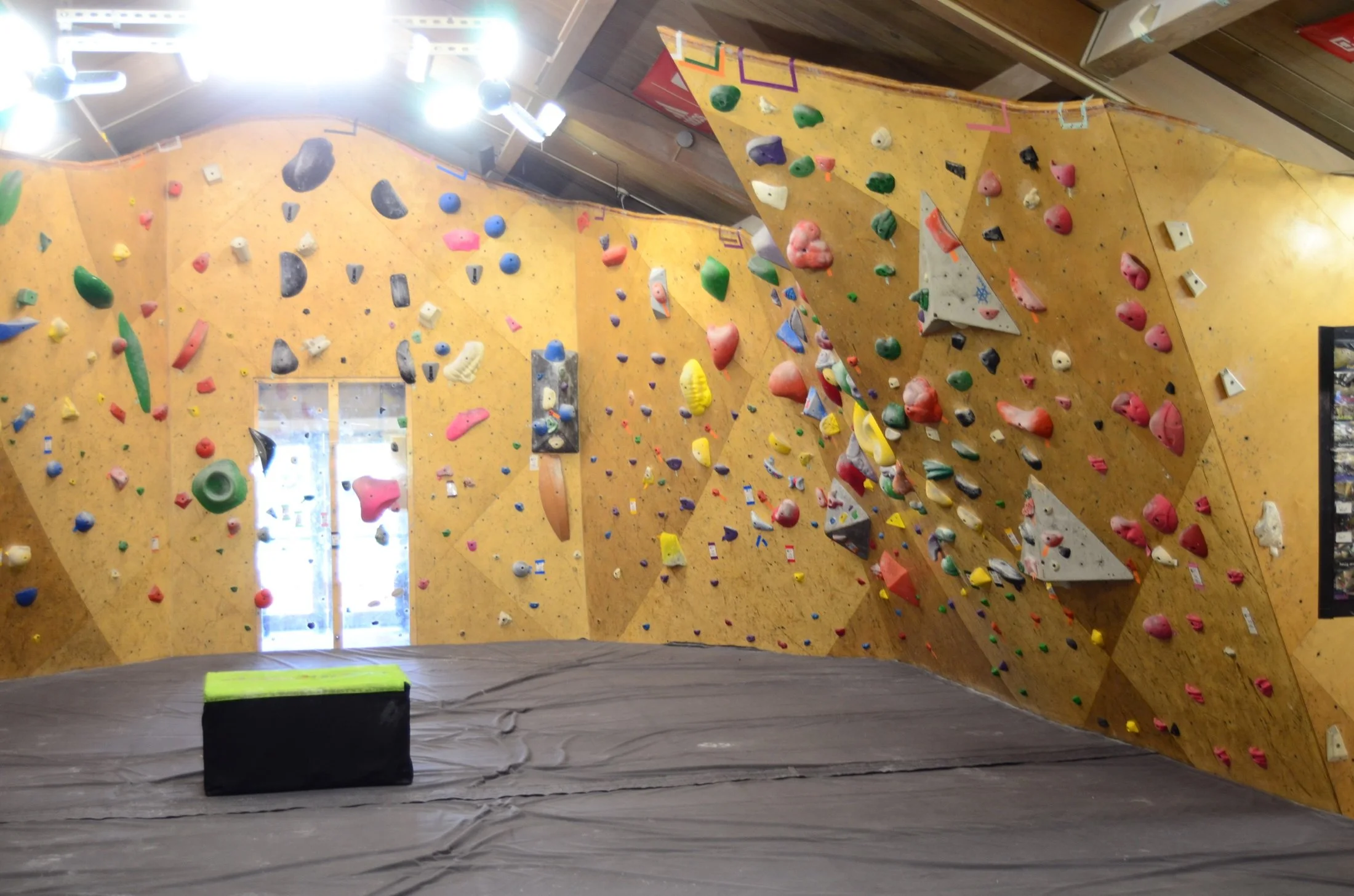 Indoor rock climbing gym with a yellow climbing wall covered in colorful holds. The wall has various overhangs and angles. There is a padded floor at the base for safety, and bright lighting is from ceiling fixtures.