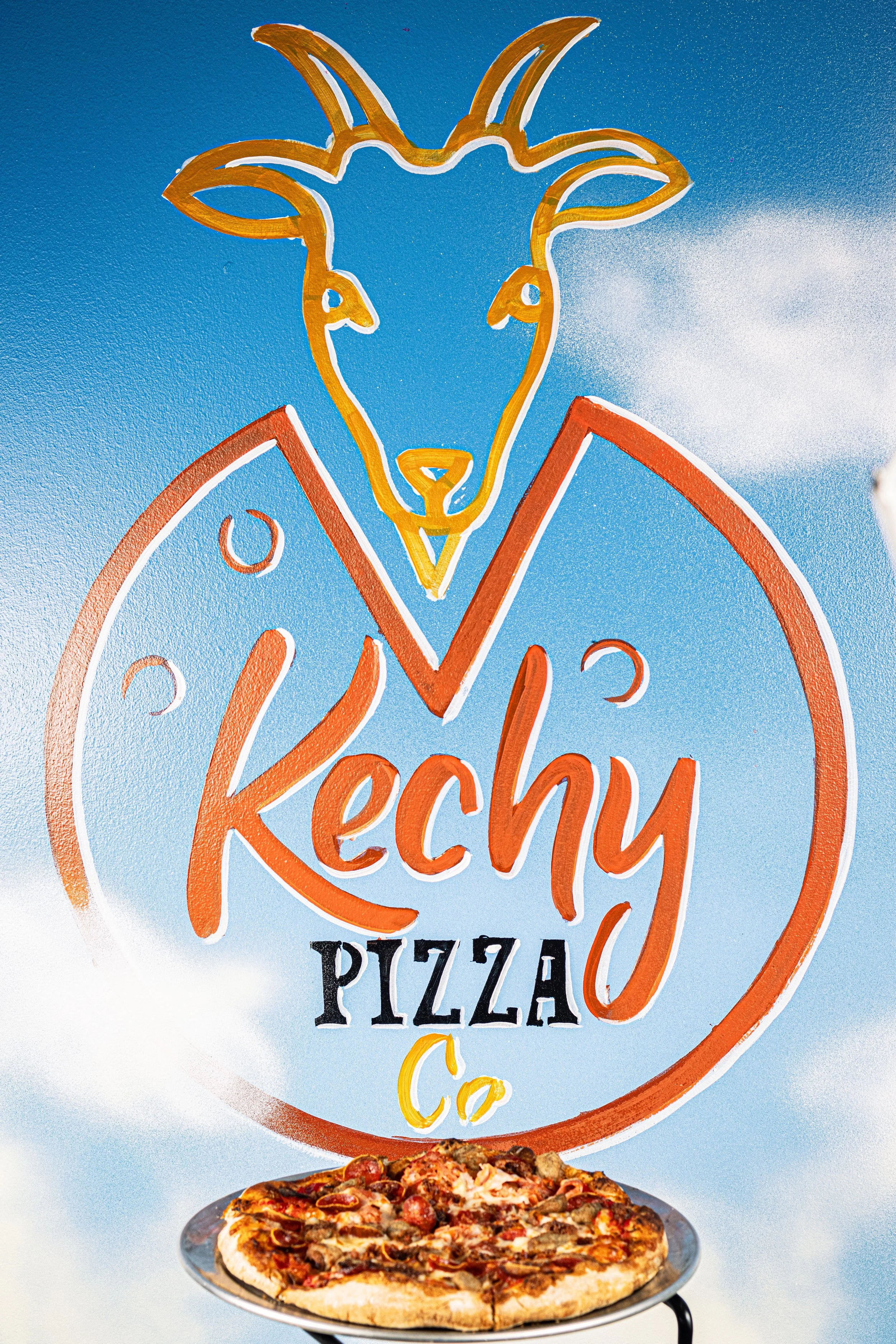 A colorful sign for Kechy Pizza Co featuring a goat's head outline with a blue background and a pizza placed below the sign. Taken at Kechy Pizza Co in downtown Baltimore