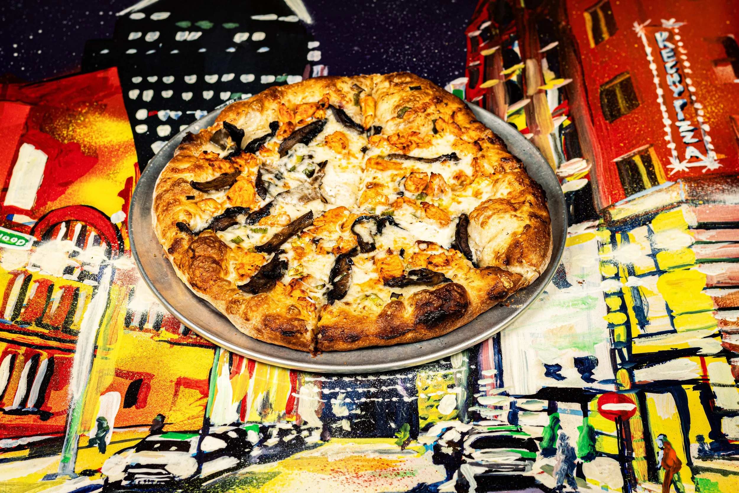 A Kechy Pizza Co Baltimroe, MD pizza with cheese, black olives, and green peppers on a metallic tray, placed on a colorful, abstract cityscape tablecloth.