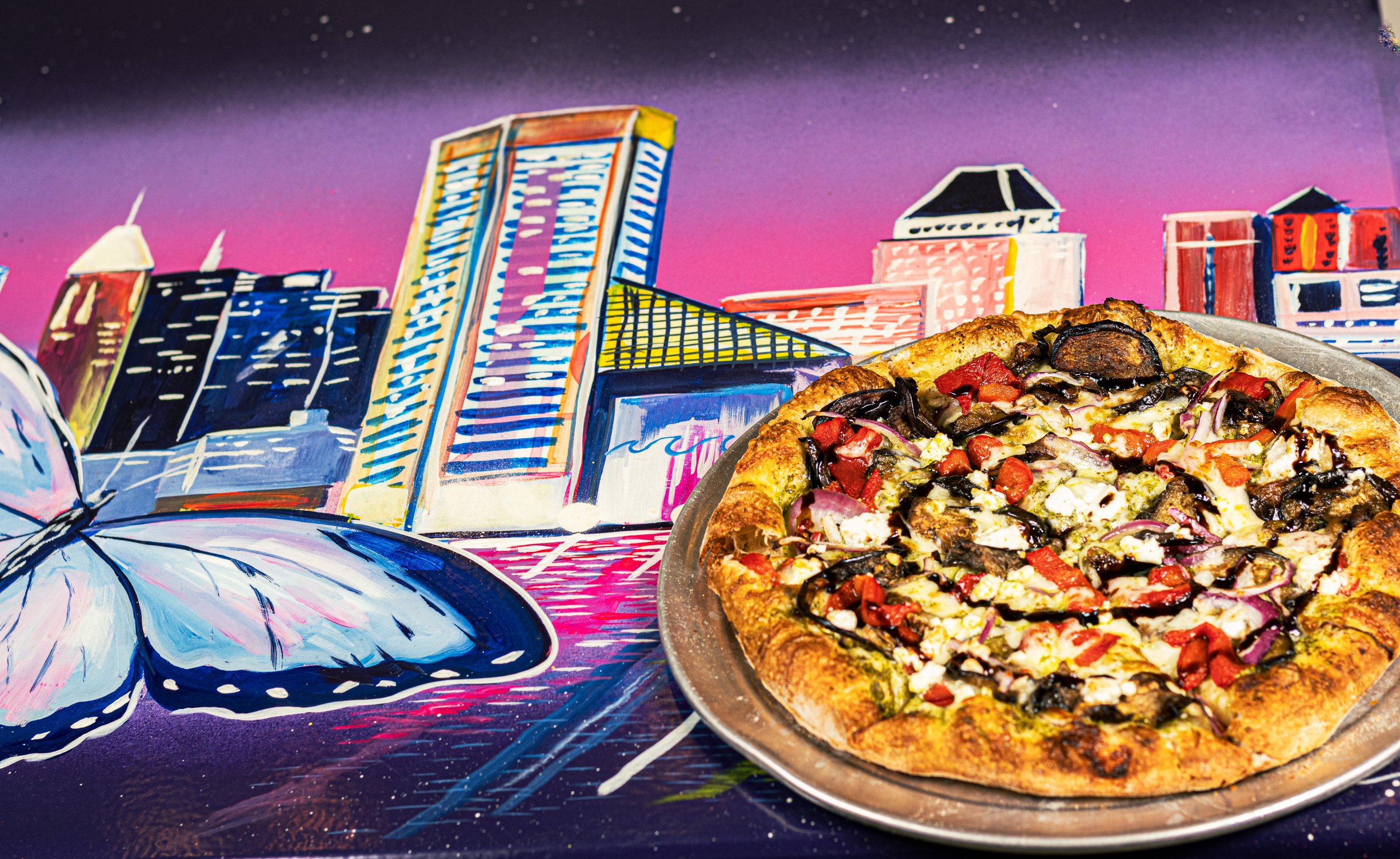 A pizza at Kechy Pizza Co with toppings including black olives, red bell peppers, red onions, and cheese on a metal tray, set against a colorful cityscape mural of Baltimore Harbor with tall buildings and a purple-pink sky.