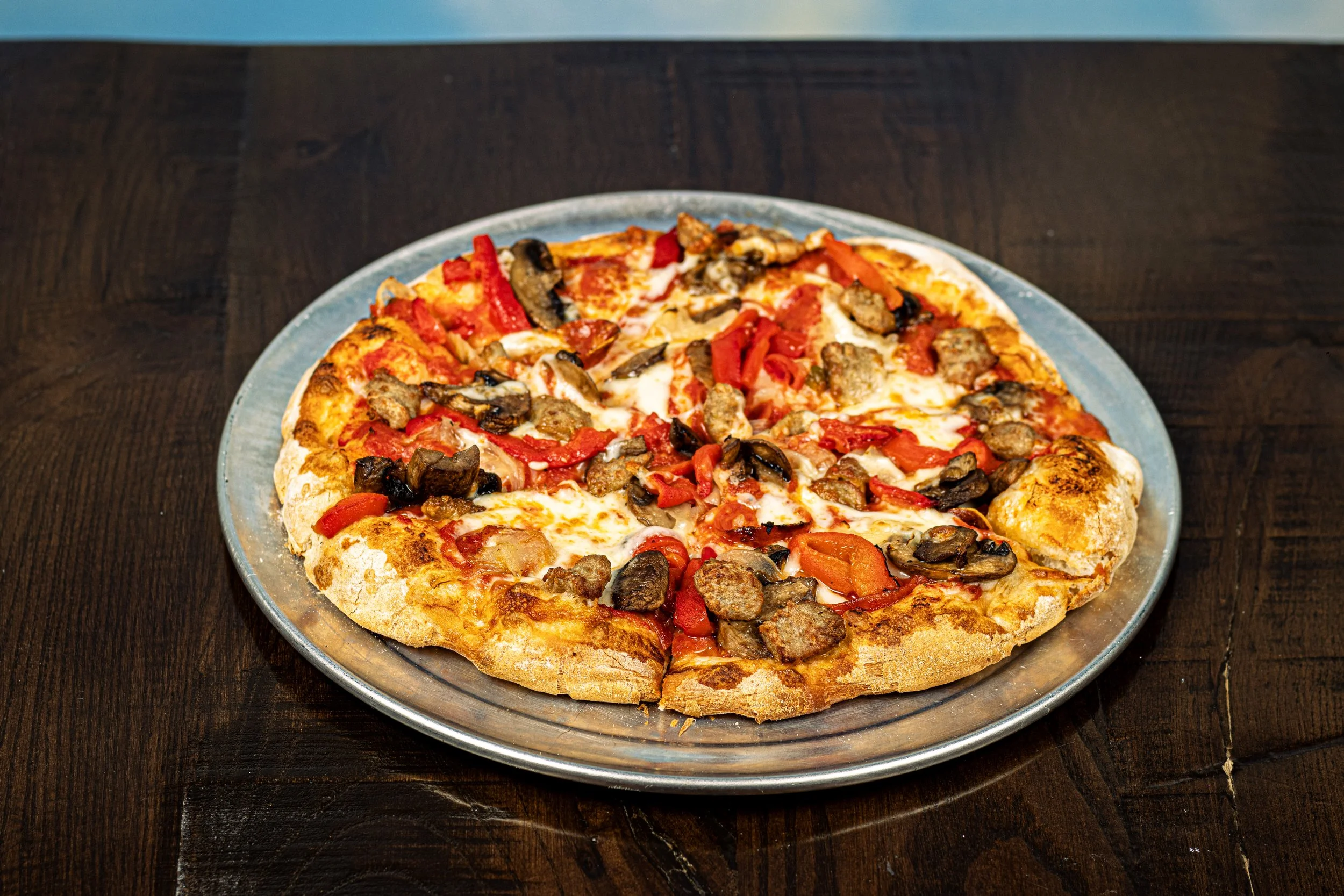 A Kechy Pizza Co, Westminster, MD pepperoni and sausage pizza with red peppers and mushrooms on a round metal tray, on a dark wooden table.