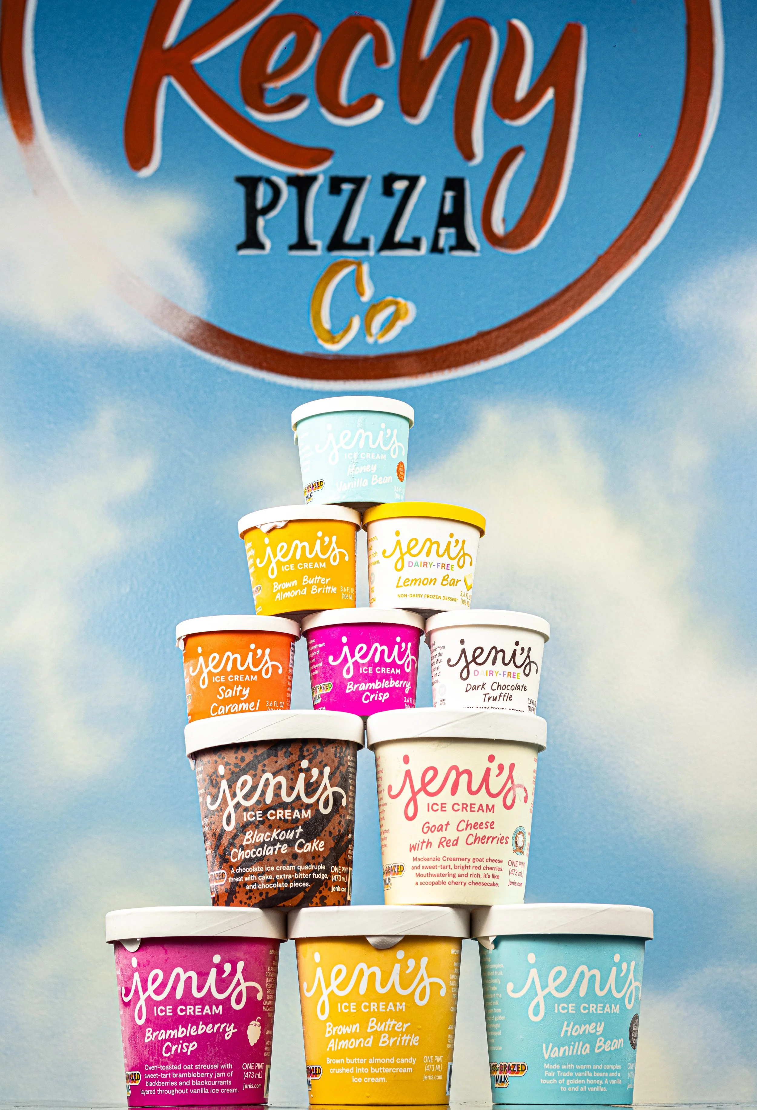 Stack of various Jeni's ice cream containers against a blue sky with clouds background, with a large Kechy Pizza Co logo on the wall behind.