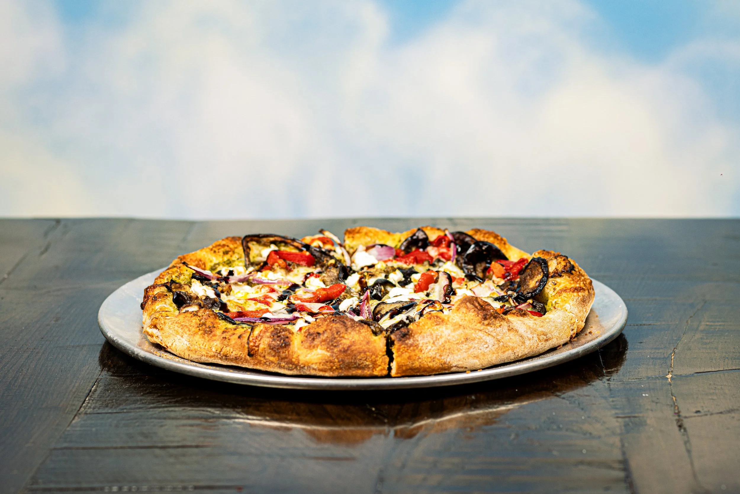 A freshly baked Kechy Pizza Co, Westminster, MD vegetable pizza with red onions, bell peppers, black olives, and cheese on a plate, placed on a dark wooden table.
