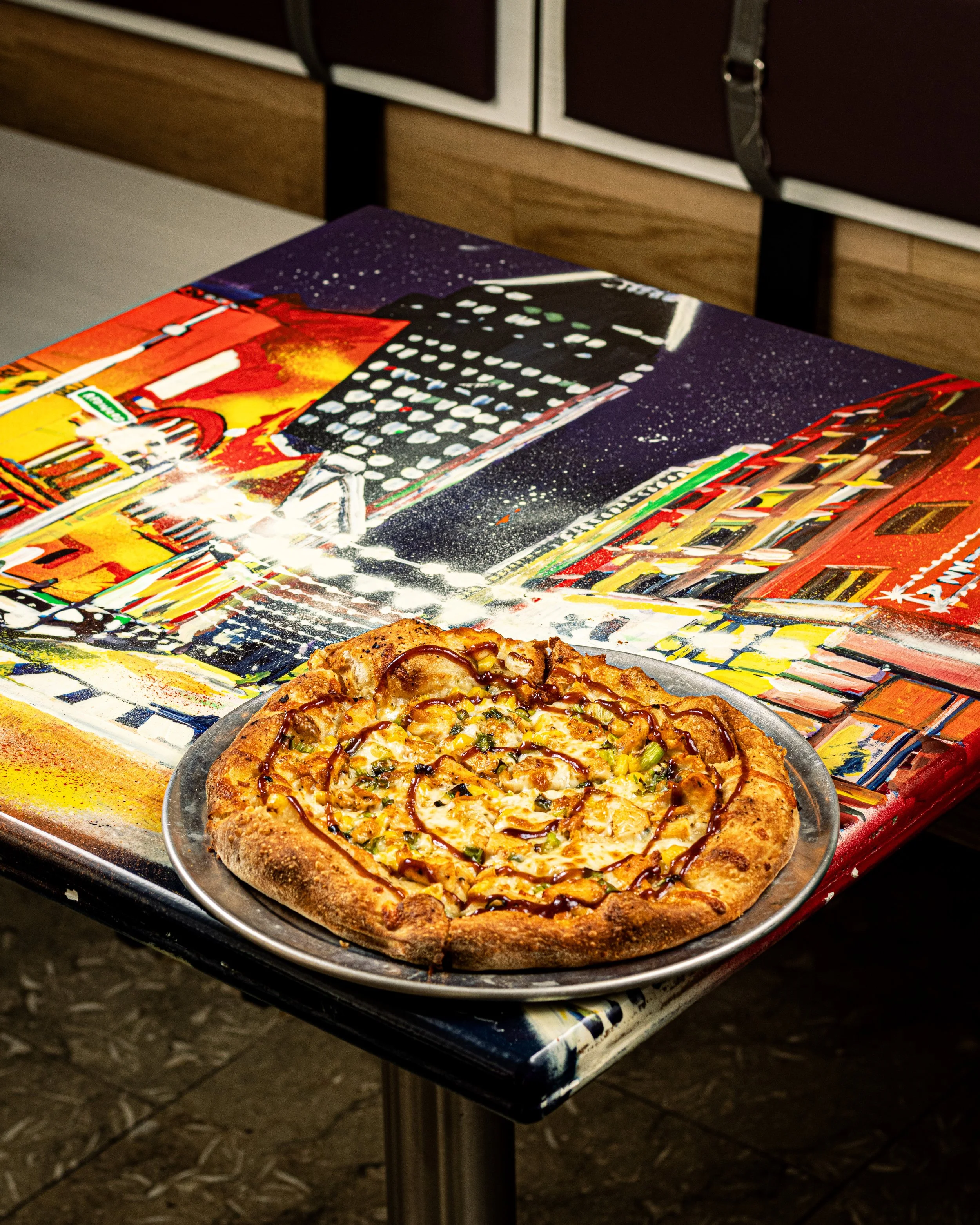 Pizza on a metal plate on a colorful table with a downtown Baltimore city scene painted on it, inside Kechy Pizza Co restaurant in downtown baltimore.