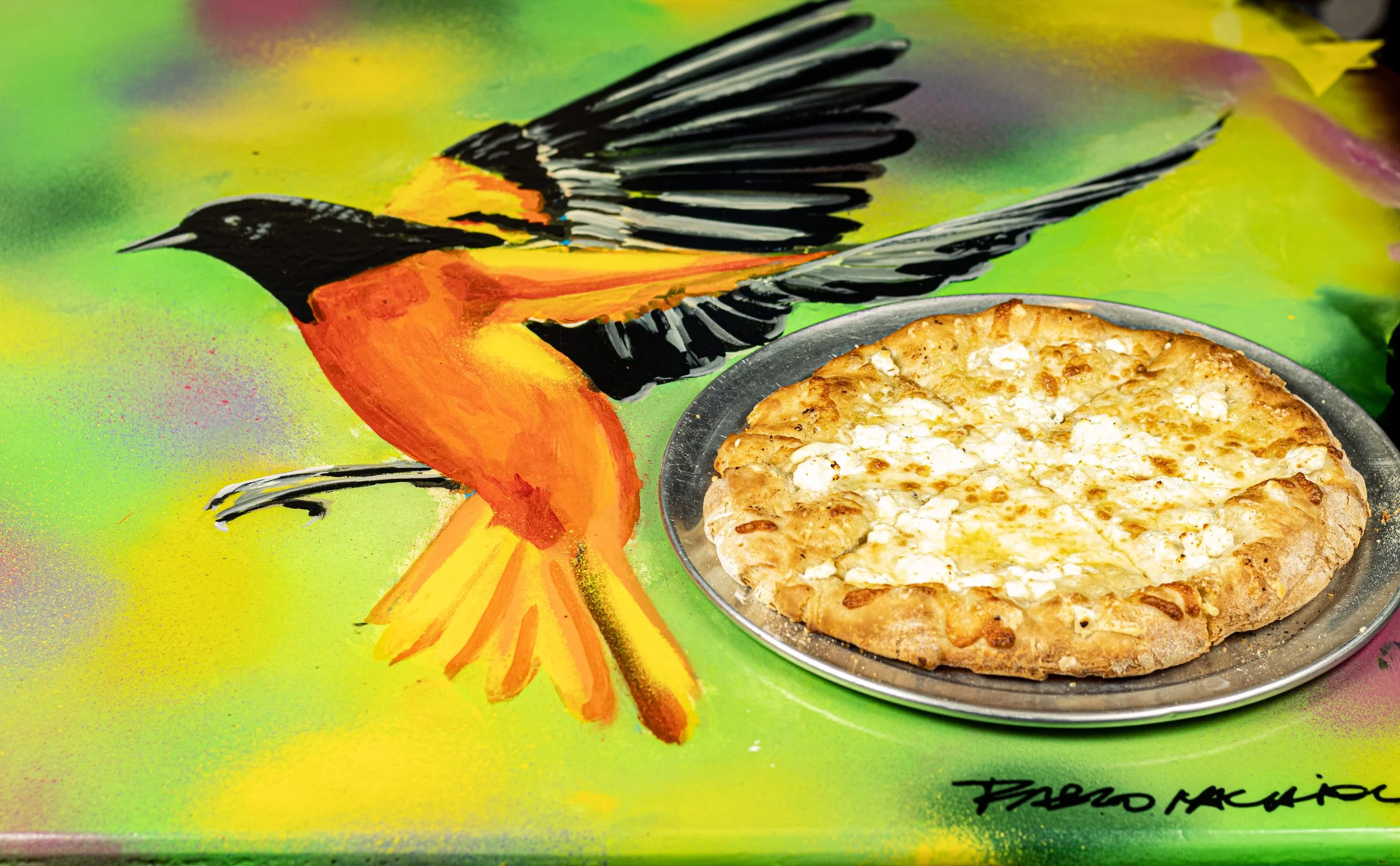 A painted mural of a Baltimore Oriole bird on a green background, with a cheese pizza on a silver tray in front of it in downtown Baltimore at Kechy Pizza Co.