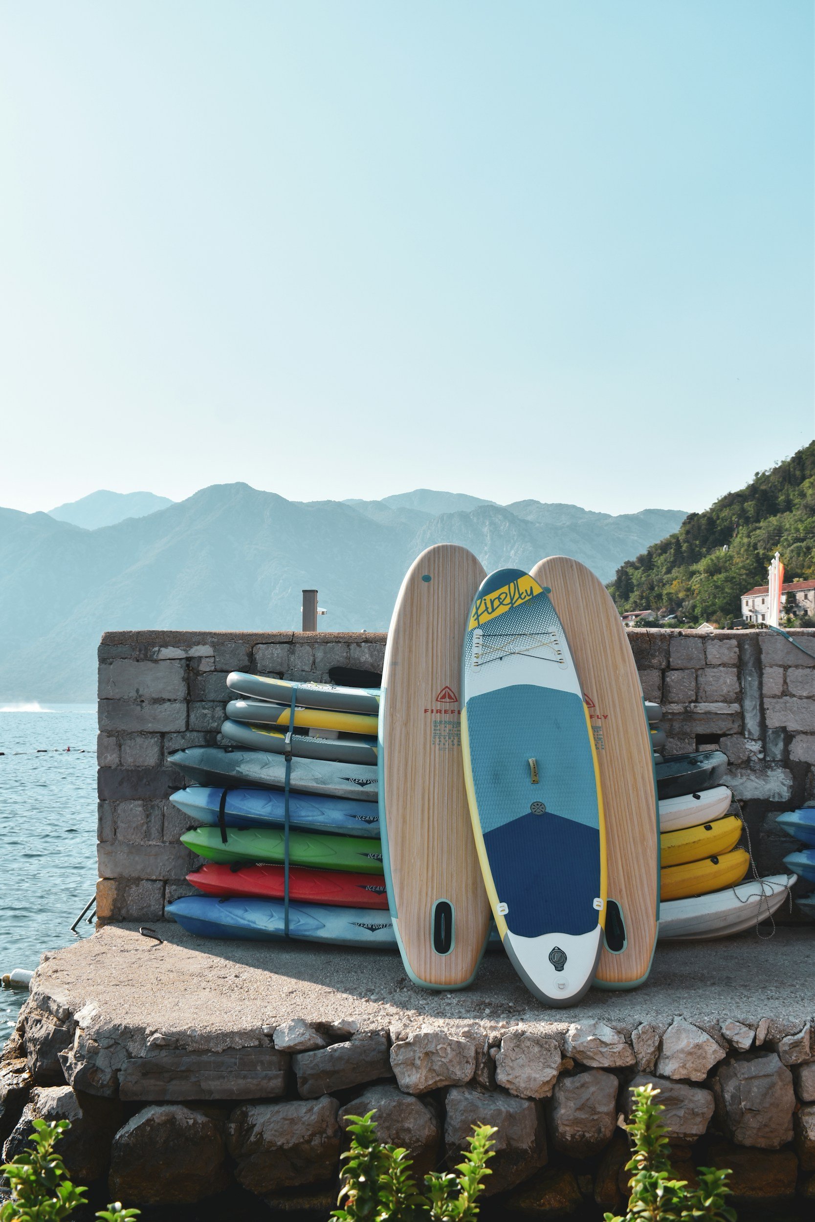Une rangée de planches de paddleboards colorés empilées contre un mur en pierre, avec deux autres paddleboards devant, l'un en bois avec des détails bleus et jaunes, l'autre en blanc avec des détails noirs, au bord de l'eau avec des montagnes en arrière-plan.