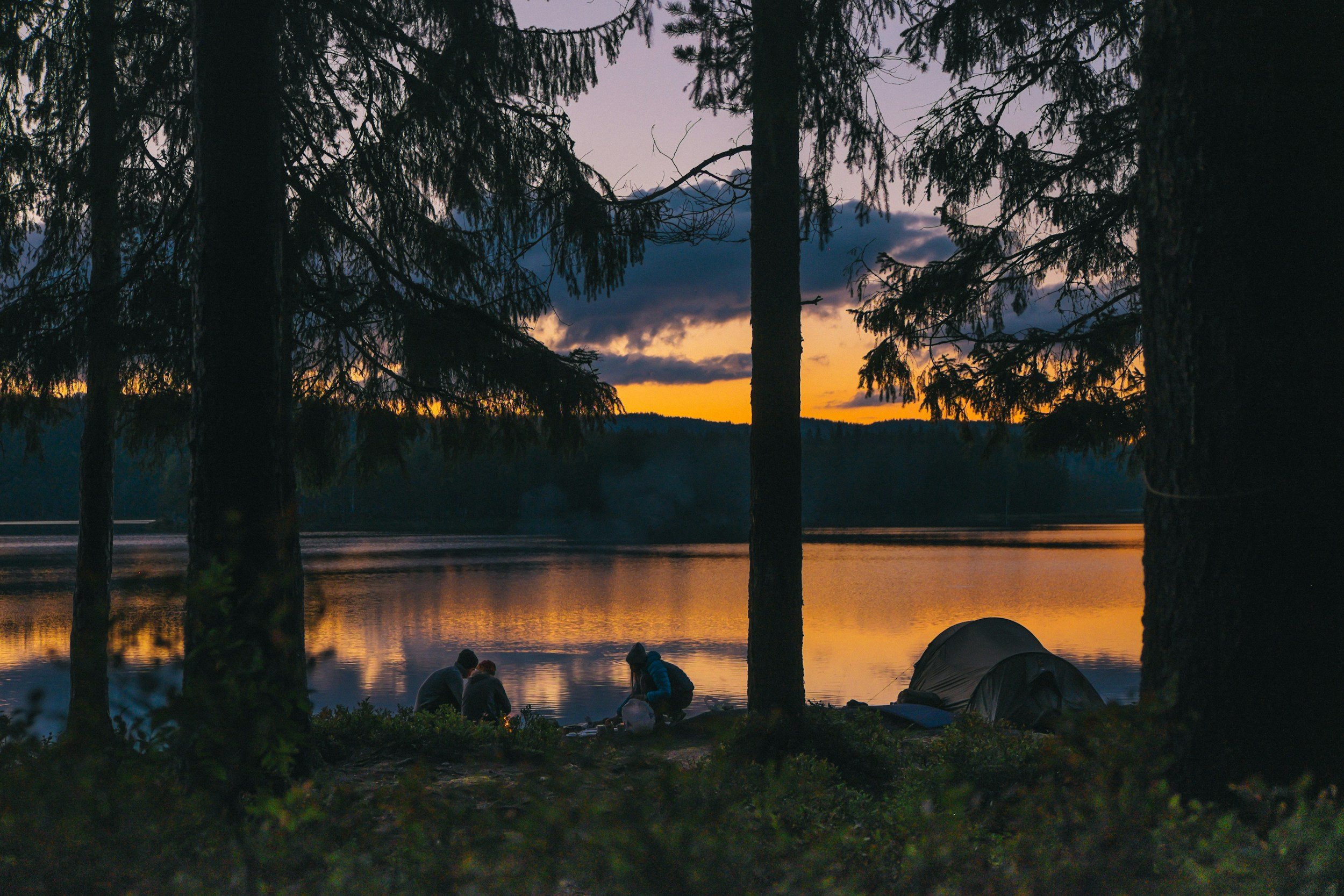 Coucher de soleil sur un lac avec des arbres en premier plan et des personnes près de l'eau.