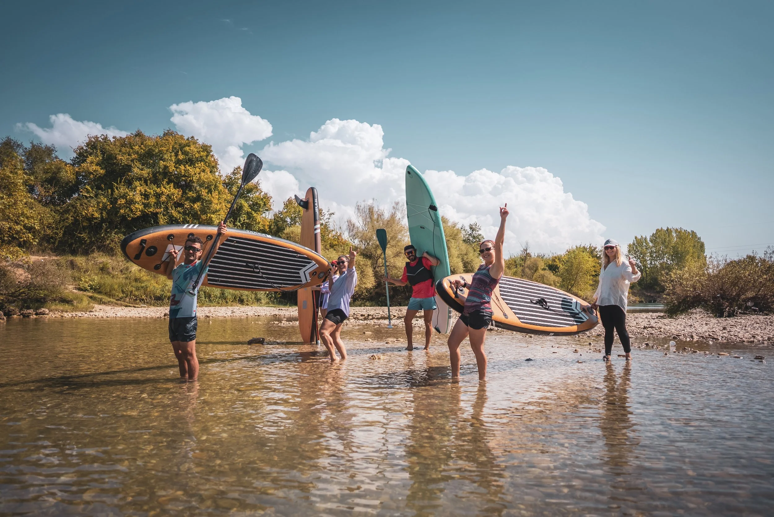 Groupe de personnes souriantes portant des planches de surf dans une rivière sous un ciel bleu avec des nuages.