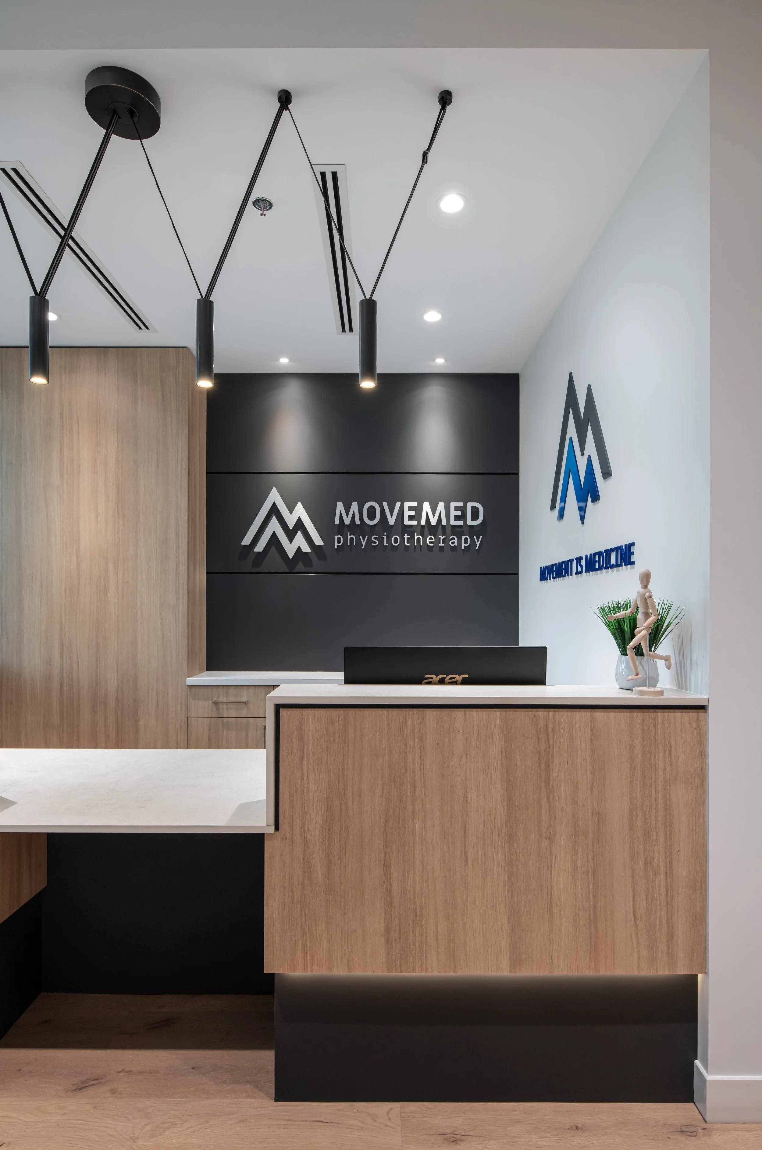 Reception desk with a black sign reading 'MOVEMED physiotherapy' and a logo, with a small sculpture and plants on the right side.