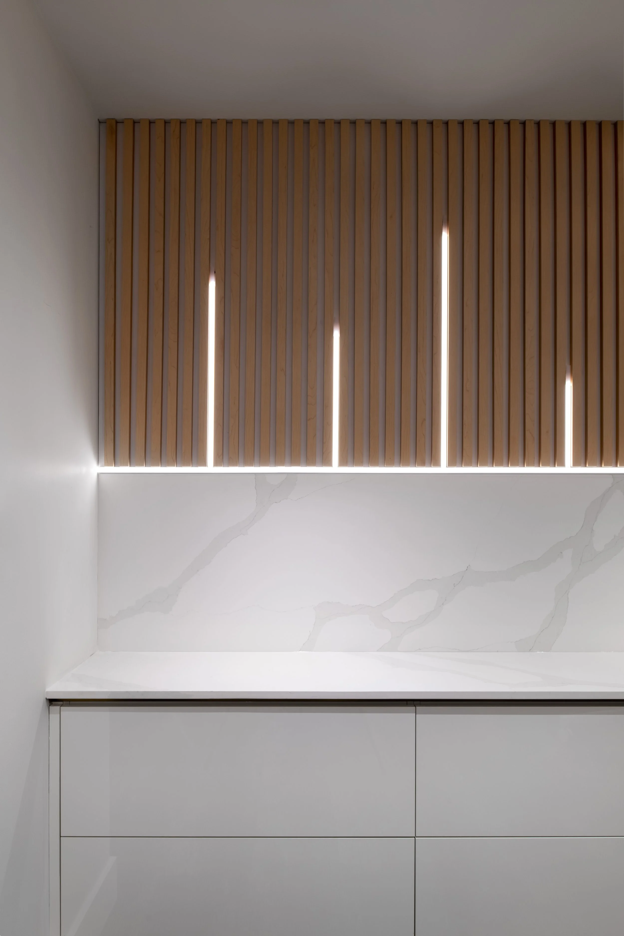Modern kitchen with white cabinets, marble backsplash, and wooden slatted wall with vertical LED lights.