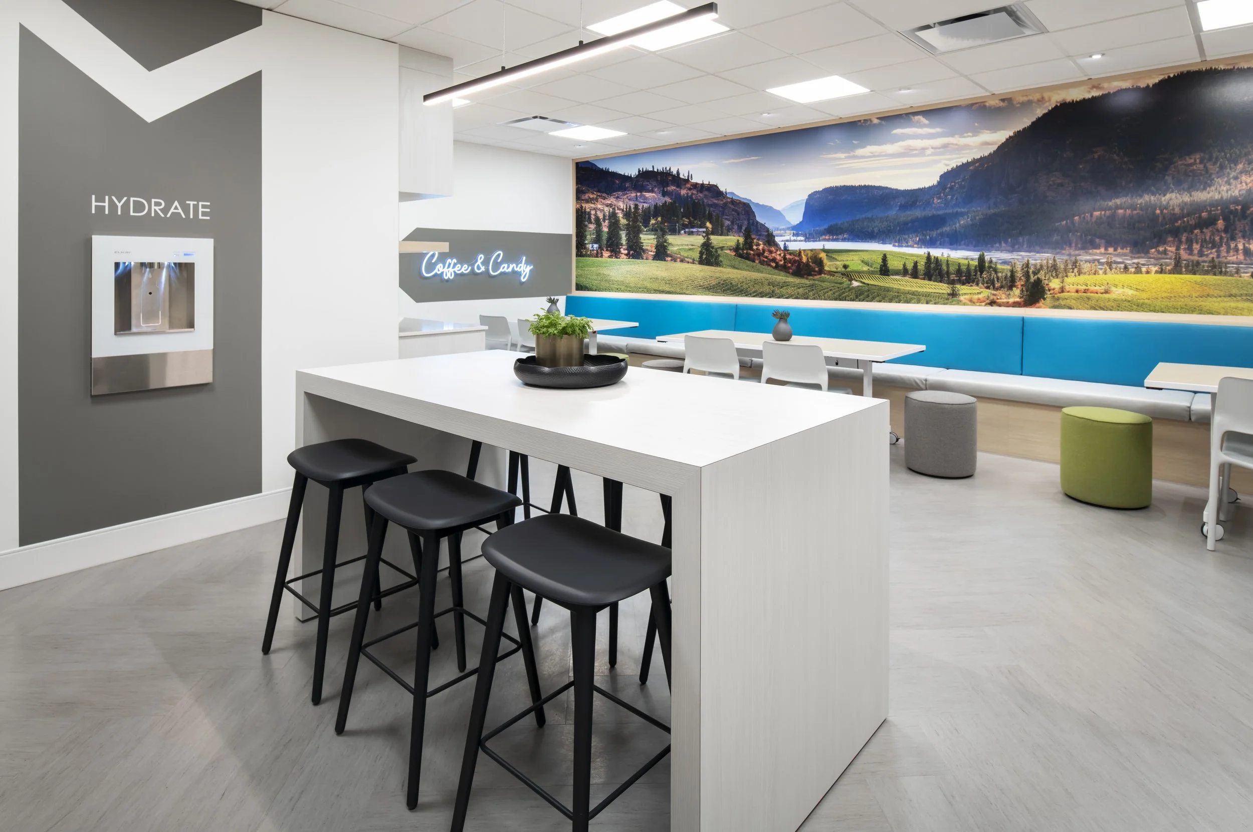 Modern lounge area with a large mural of a mountain landscape, white tables and chairs, a blue bench along the wall, and a high table with black stools in the foreground.