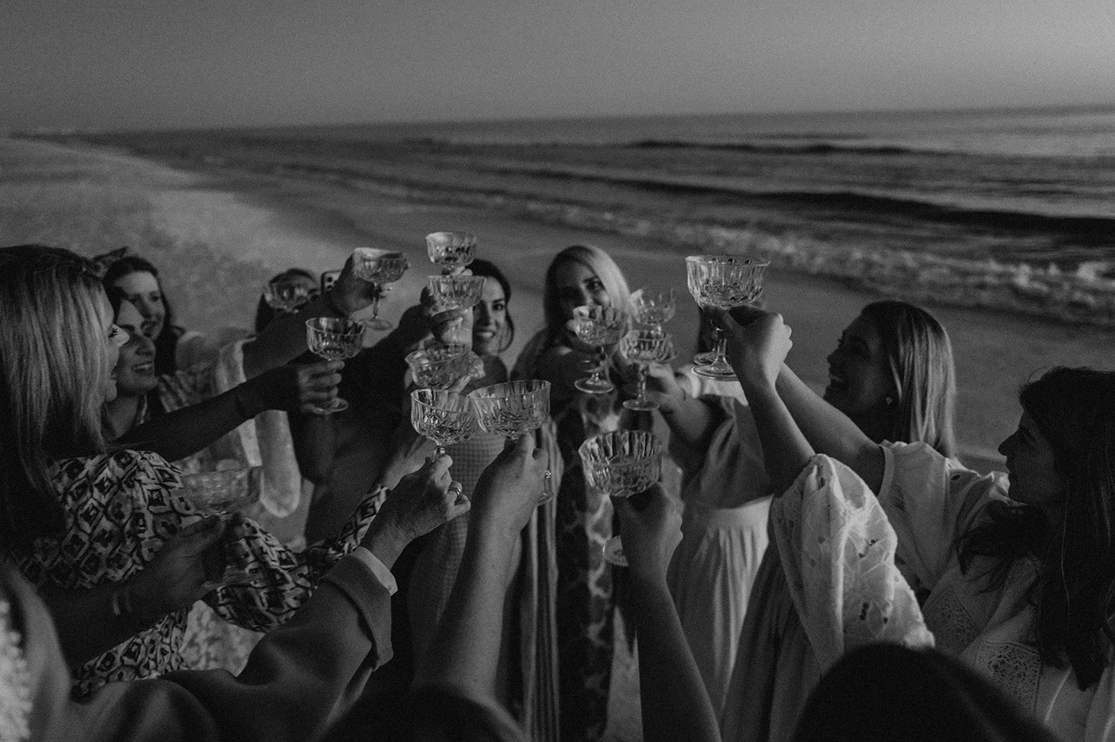 Group of women in dresses celebrating on a beach at night, raising glasses in a toast.