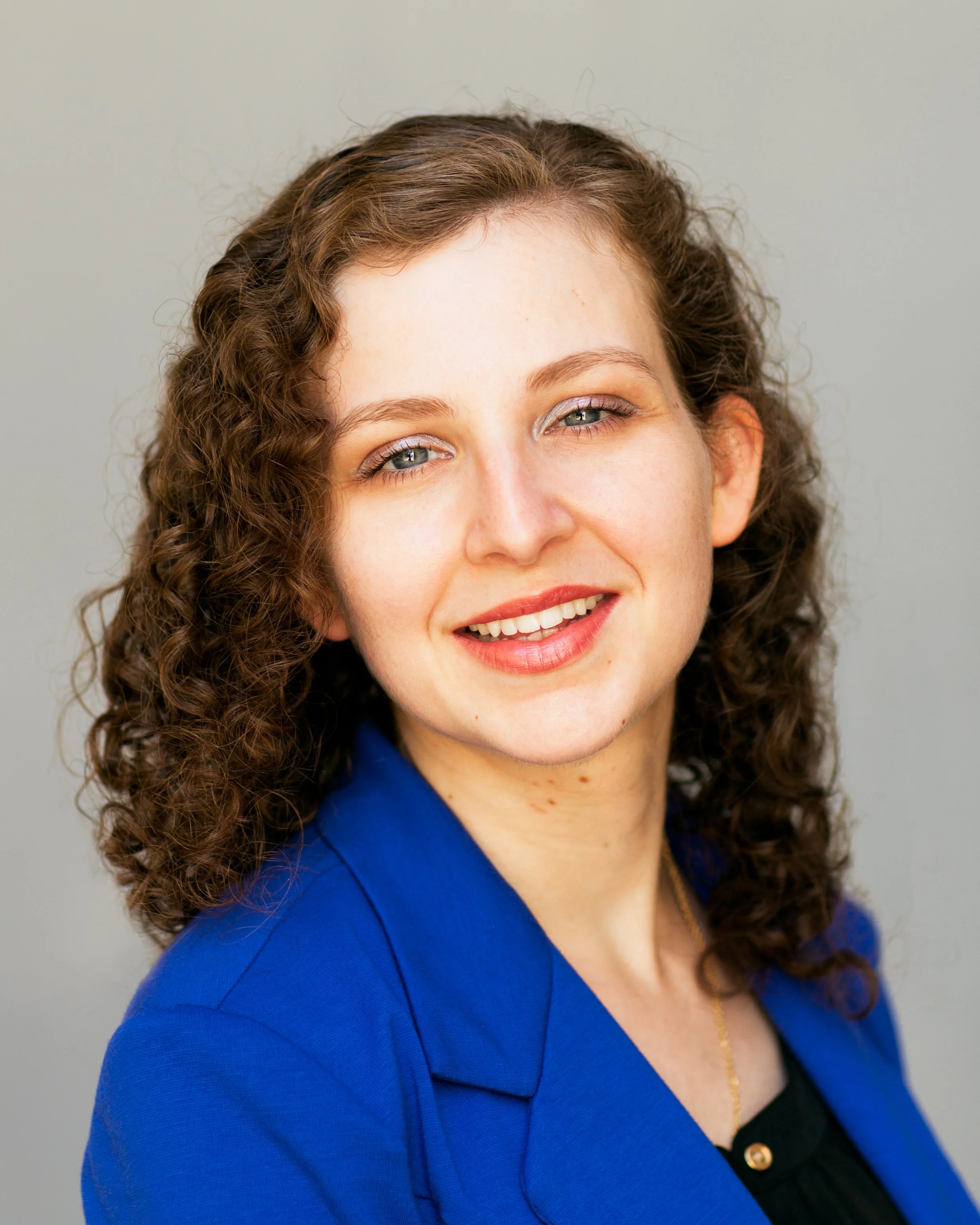 Portrait of a woman with curly brown hair, wearing a blue blazer and a gold necklace, smiling softly against a neutral background.