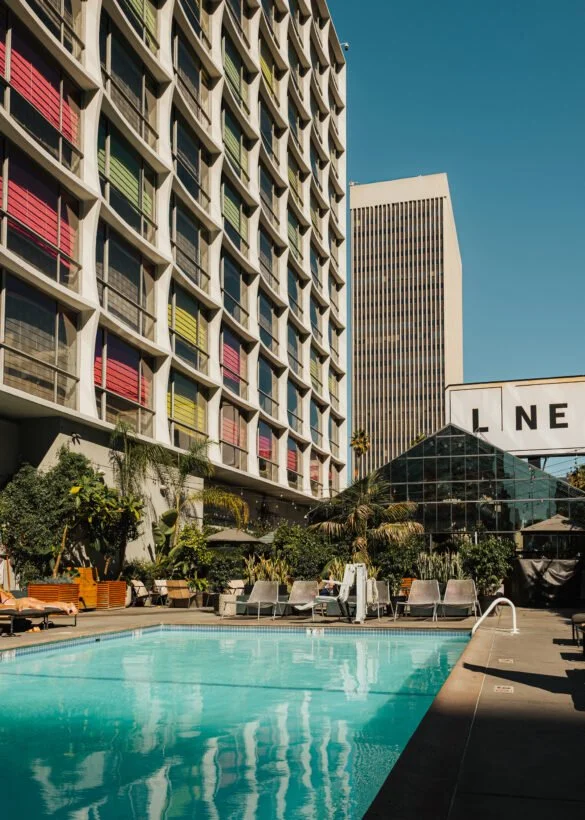 Poolside area with lounge chairs and plants, high-rise buildings in the background, and a geometric glass structure at a hotel or apartment complex.