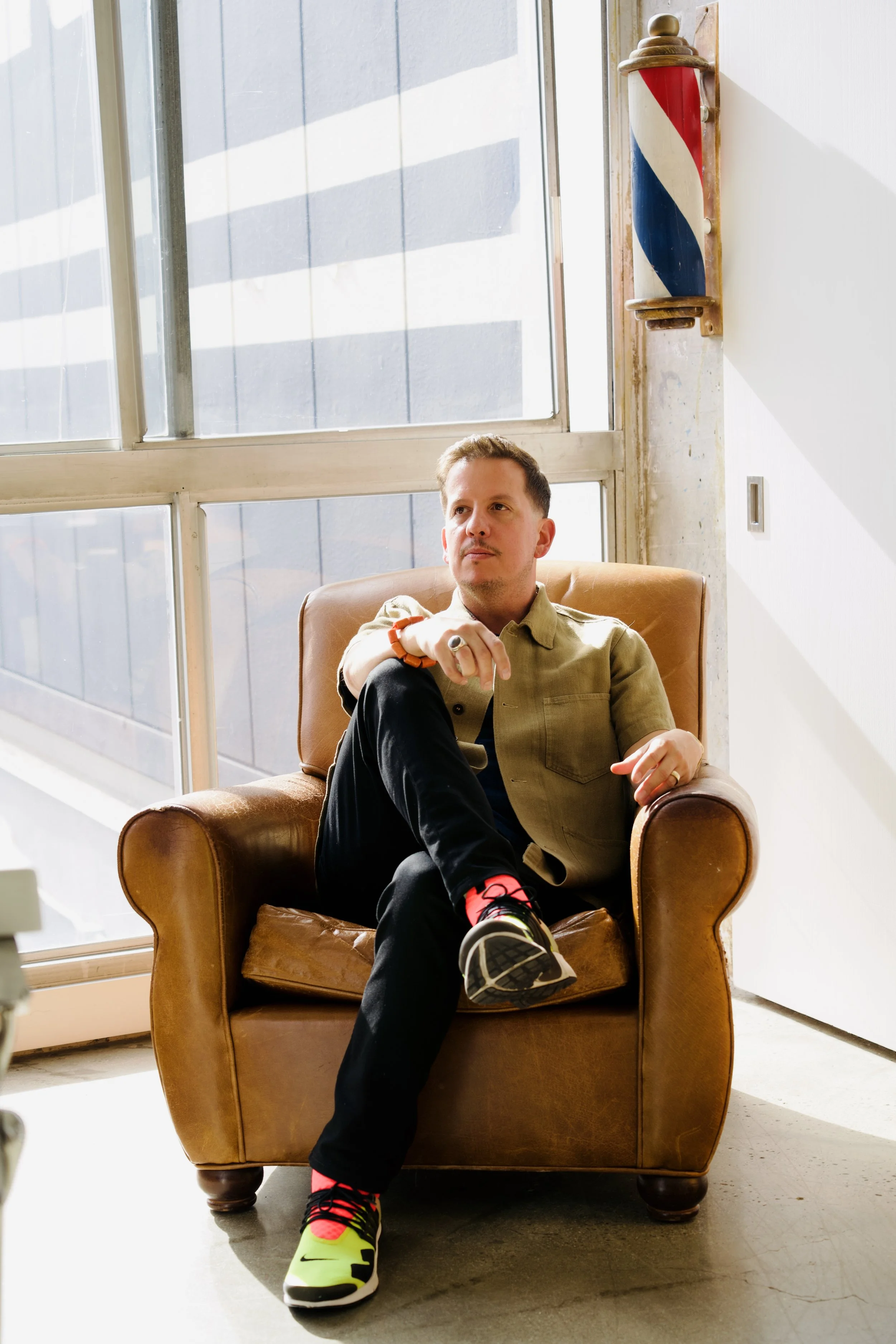 A man sitting in a brown leather armchair near a large window with sunlight, a barber pole hangs on the wall behind him.