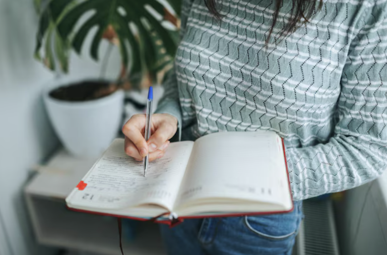 Person writing in a planner with a pen, wearing a gray patterned shirt, with a potted plant in the background.