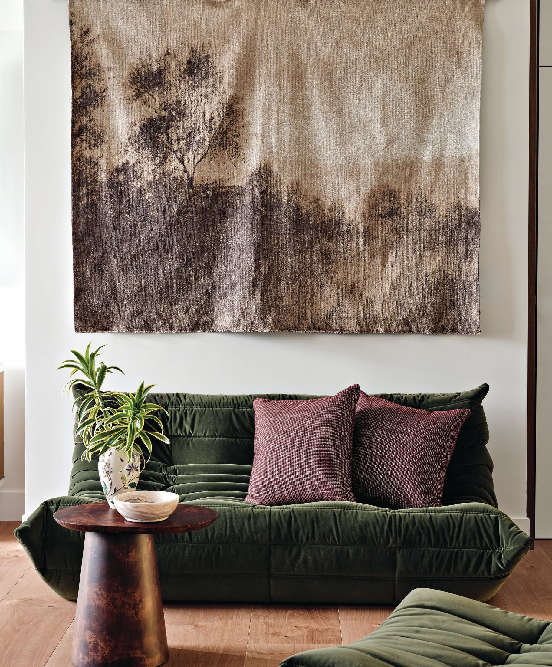 A living room with a dark green velvet sofa, two mauve throw pillows, a small wooden side table with a white ceramic bowl and a plant, and an abstract landscape artwork on the wall above the sofa.
