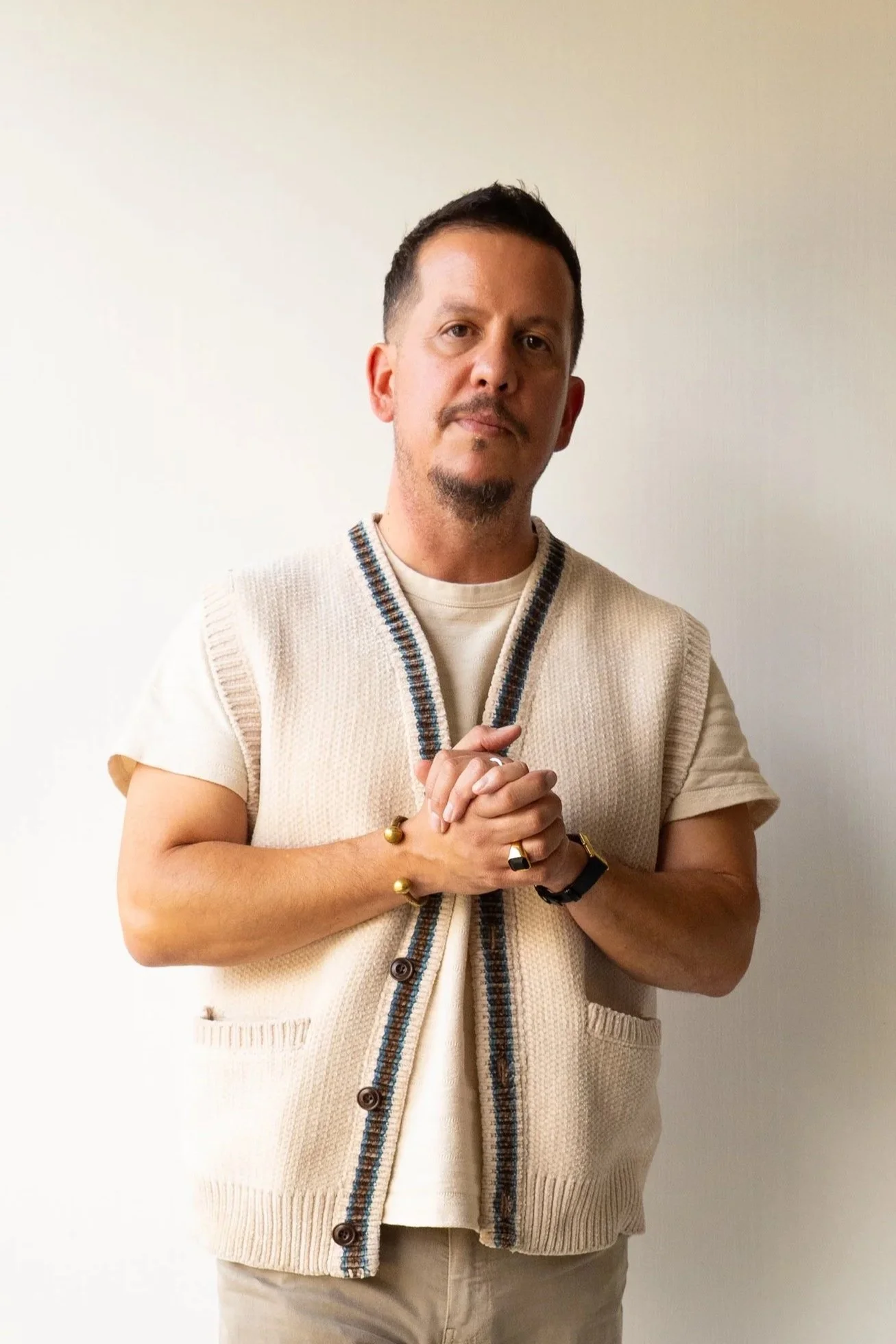 Man with short dark hair and facial hair, wearing a beige knit vest over a cream T-shirt, standing against a white background with hands clasped