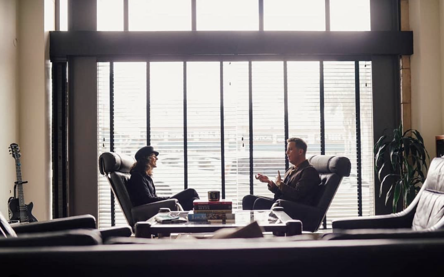Two people sitting in office chairs having a conversation in a modern office with large window blinds, a guitar leaning against the wall, and a plant in the corner.