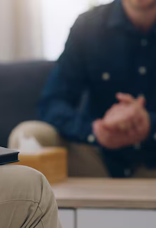 A person in a dark blue shirt sitting across a table, with hands clasped. There is a wooden box and other objects on the table. The background appears to be an indoor setting.