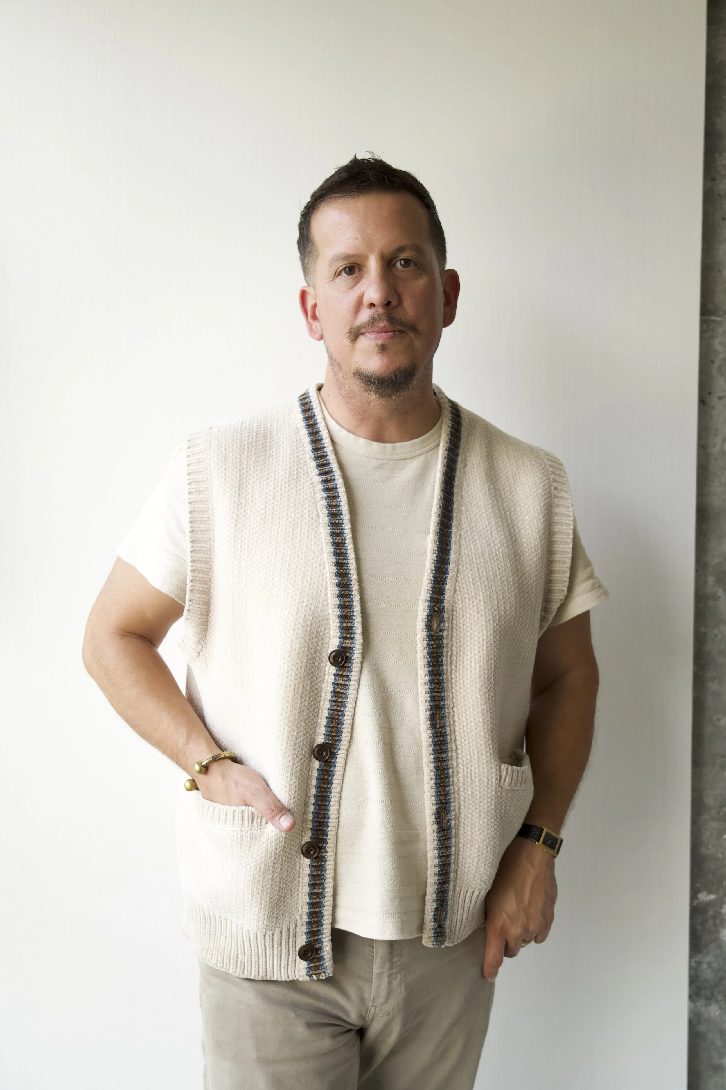 Man posing with hands in pockets, wearing beige vest over cream shirt, standing against plain white wall.