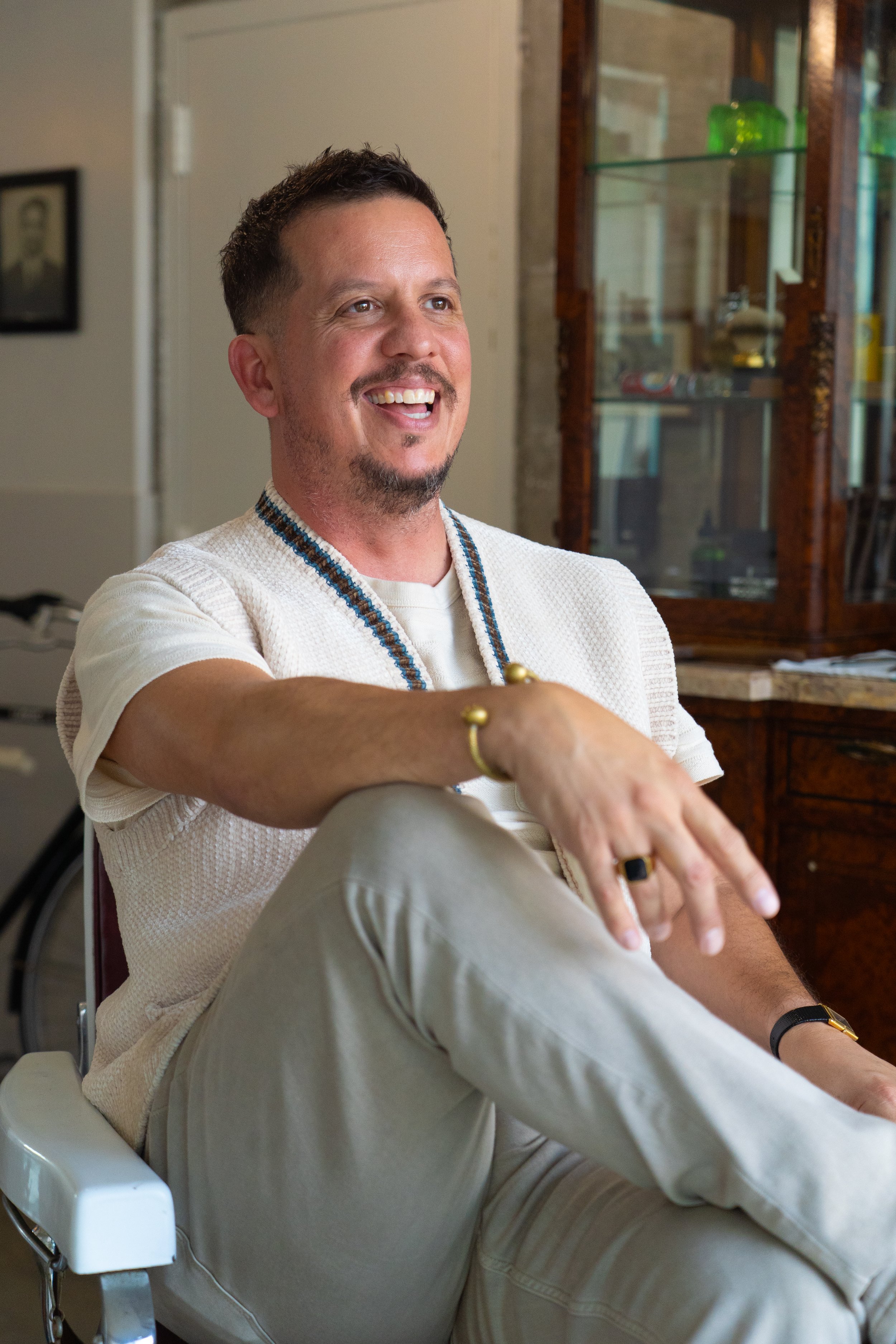 A smiling man with short dark hair, wearing a white textured shirt with a beaded necklace, sitting in a chair inside a room with wood and glass display cabinets.