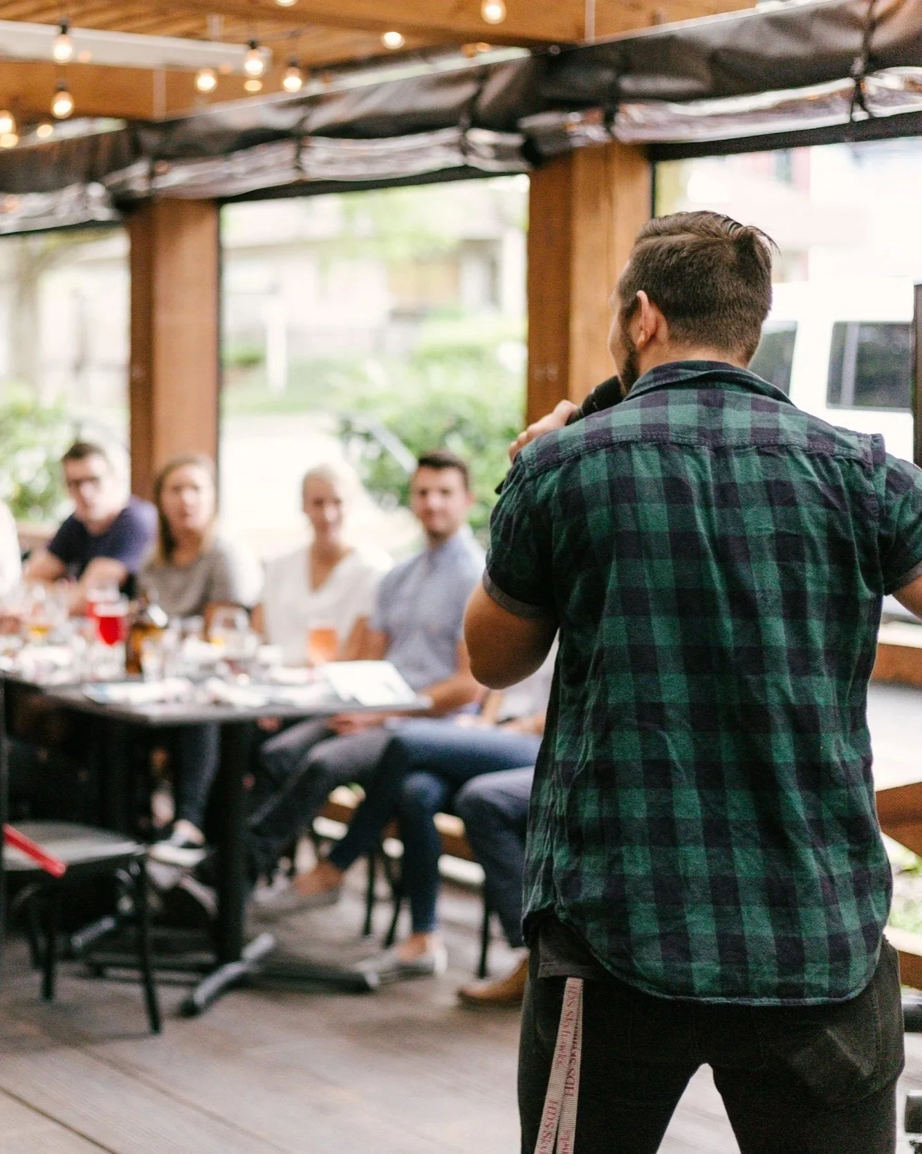 A man with a beard and a plaid shirt speaking into a microphone to a seated audience in a restaurant or cafe with large windows and wooden decor.